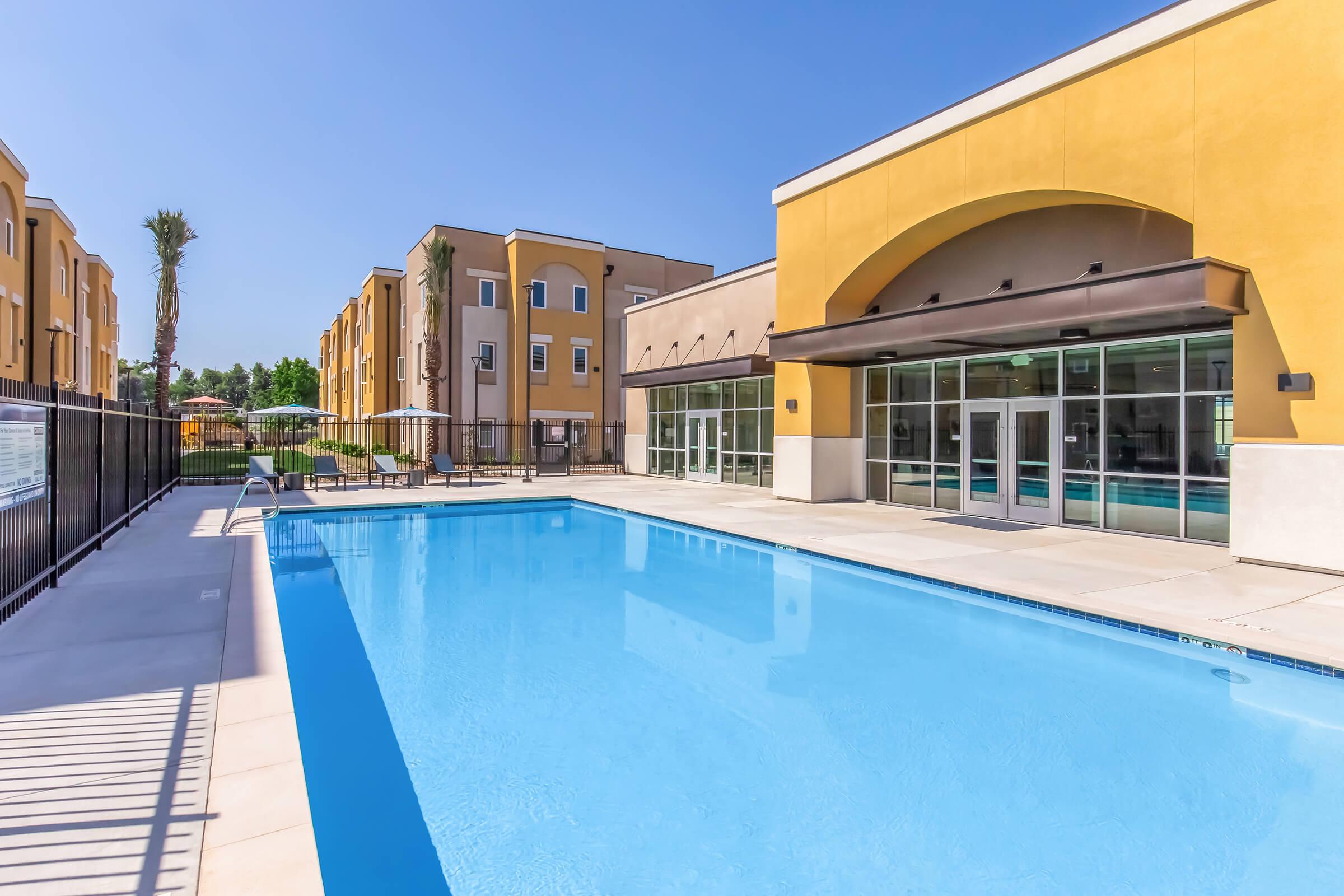 A clear blue swimming pool surrounded by a patio area, with lounge chairs and umbrellas. In the background, modern yellow and beige apartment buildings are visible under a bright blue sky. The scene conveys a welcoming and relaxing atmosphere.