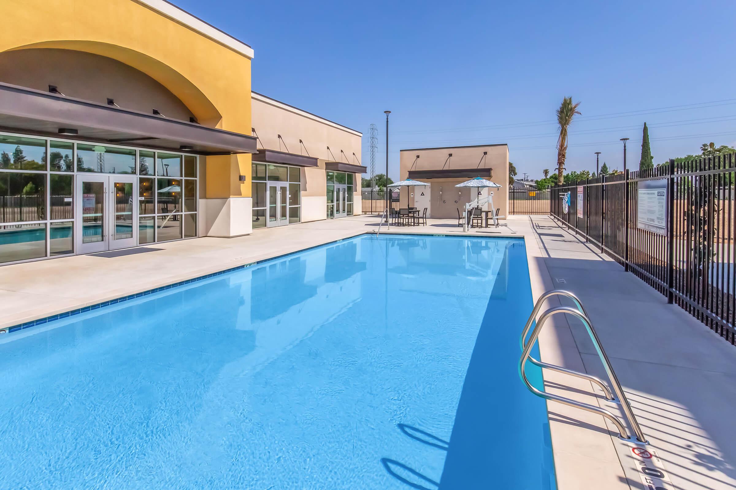 A clear blue swimming pool surrounded by a patio area, with lounge chairs and umbrellas. The pool area is enclosed by a black fence, and there are modern buildings with large windows in the background. Bright sunshine and clear blue skies enhance the scene, creating a relaxing atmosphere.