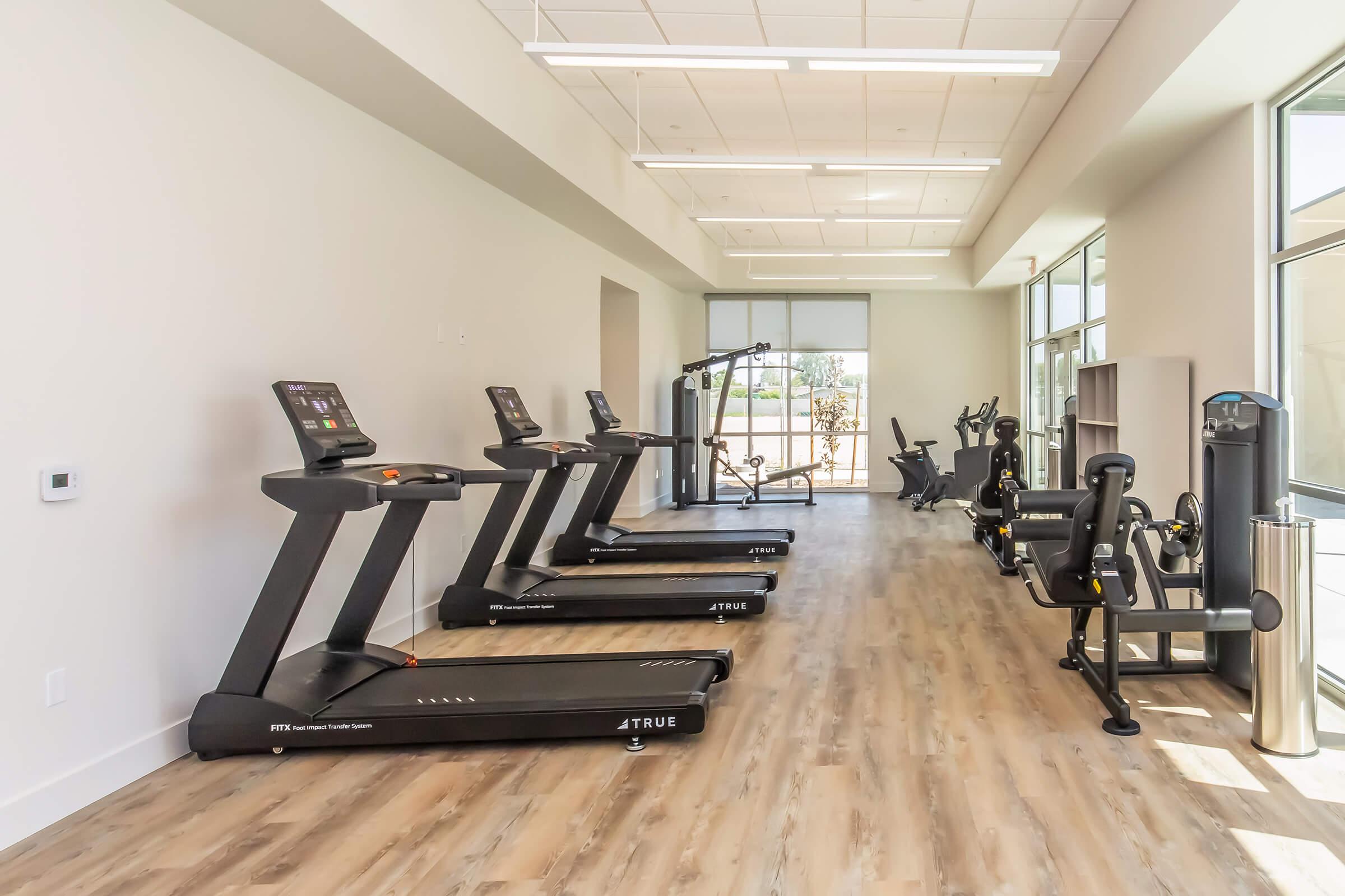 A modern gym interior featuring three treadmills lined up near large windows. Additional fitness equipment, including a stationary bike and a weight machine, is visible. The space is well-lit with wooden floors and a clean, minimalistic design.