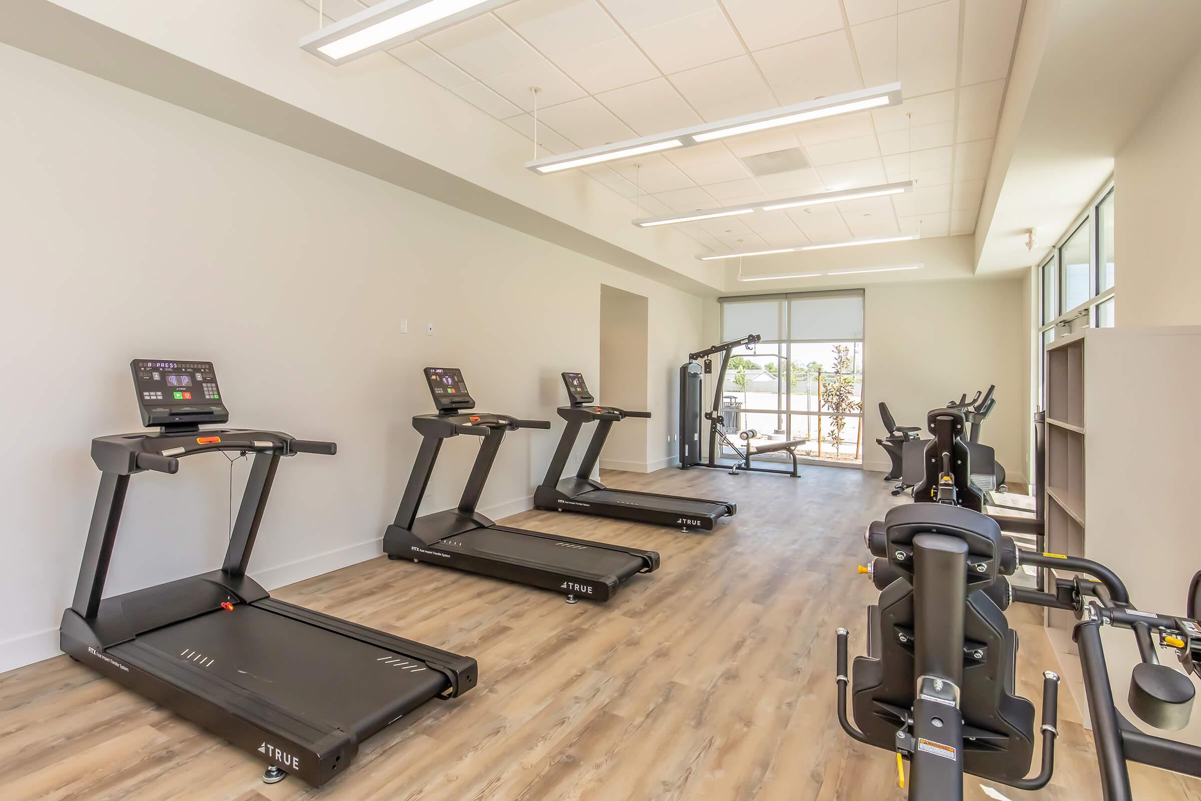 A bright and spacious gym area featuring several treadmills lined up against a wall. The floor is made of light wood, and large windows allow natural light to illuminate the space. There’s also a multi-use exercise machine in the background, contributing to a well-equipped fitness environment.