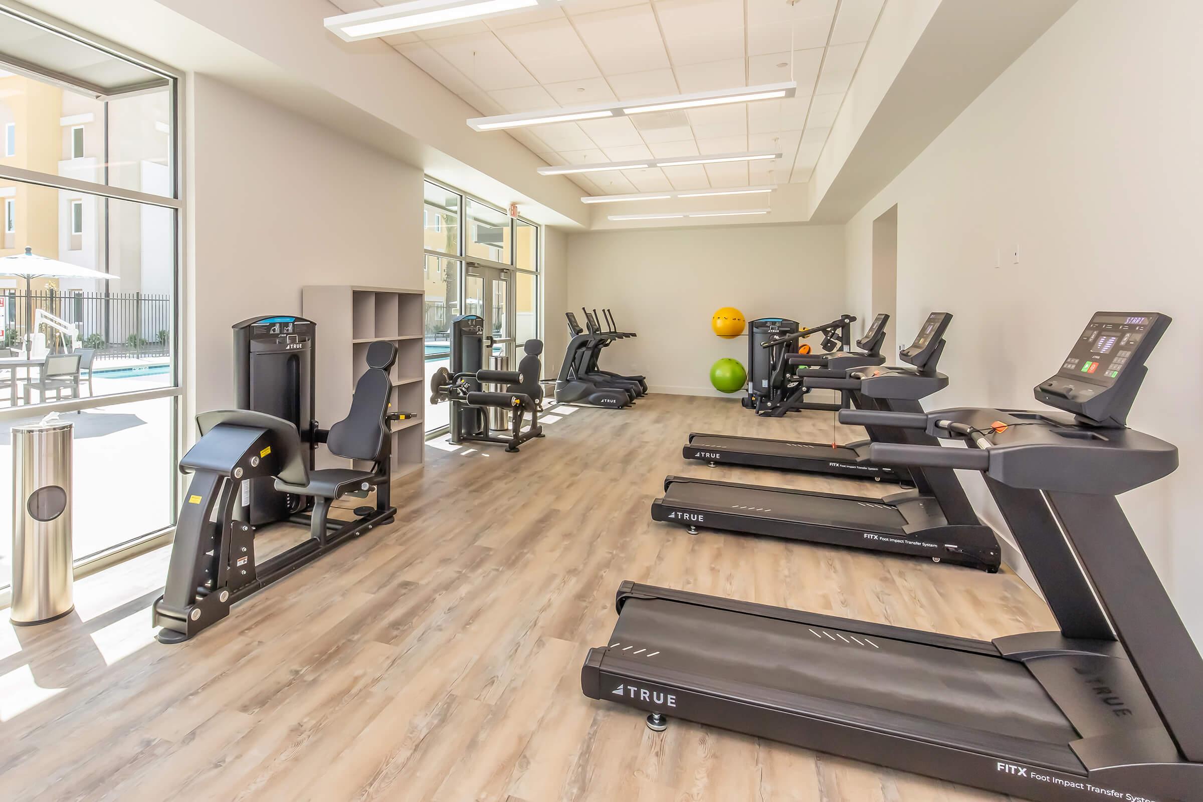 Indoor fitness area featuring multiple treadmills and exercise equipment. The space is well-lit with large windows, allowing natural light to enter, and includes exercise balls in the corner. The flooring is wooden and the overall atmosphere is modern and inviting, ideal for workouts.