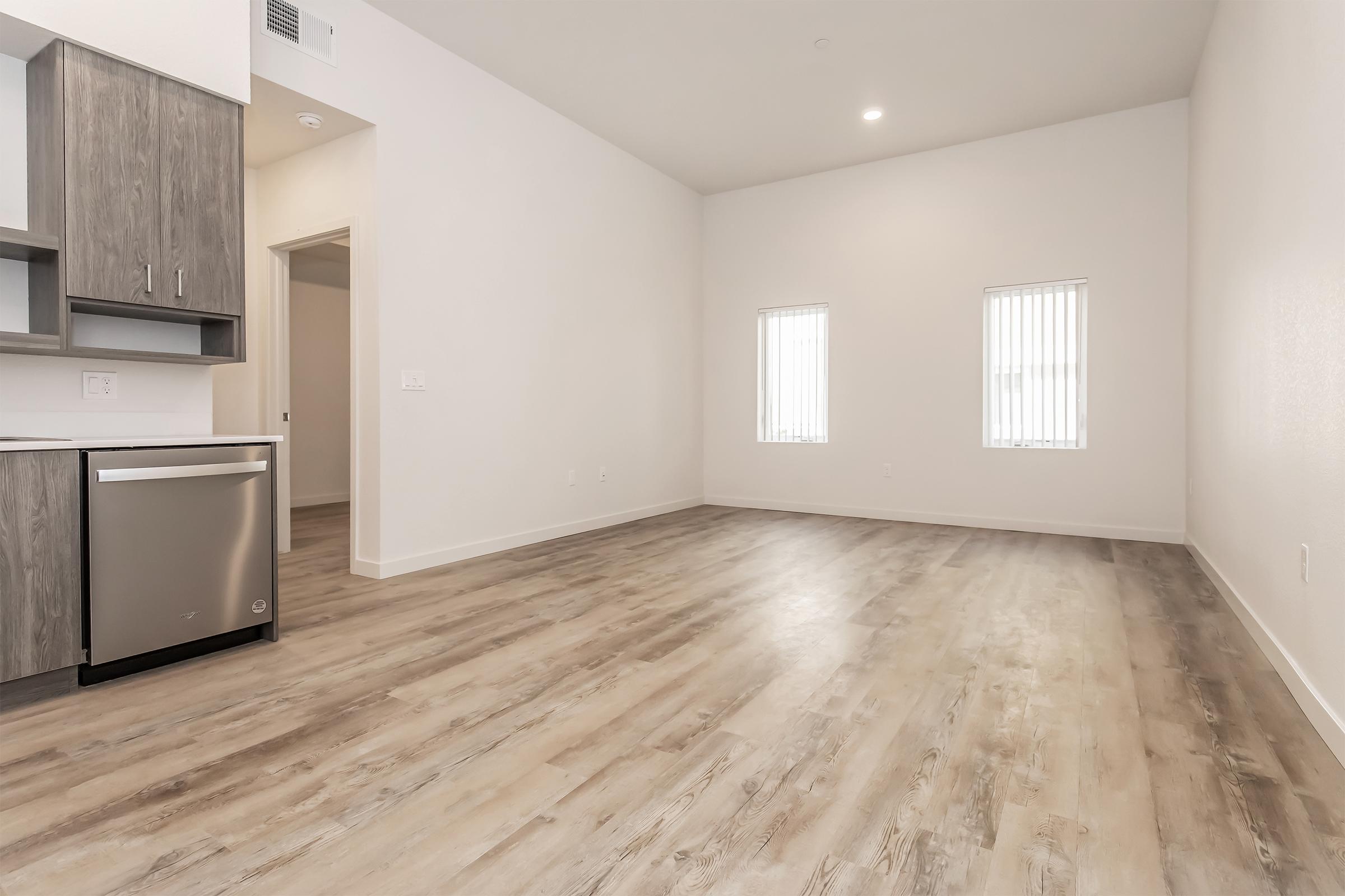 A spacious, modern living area with light-colored wood flooring. The room features a kitchen area with sleek cabinets and a dishwasher, and two vertical window treatments allowing natural light. The walls are painted white, creating a bright and airy atmosphere.