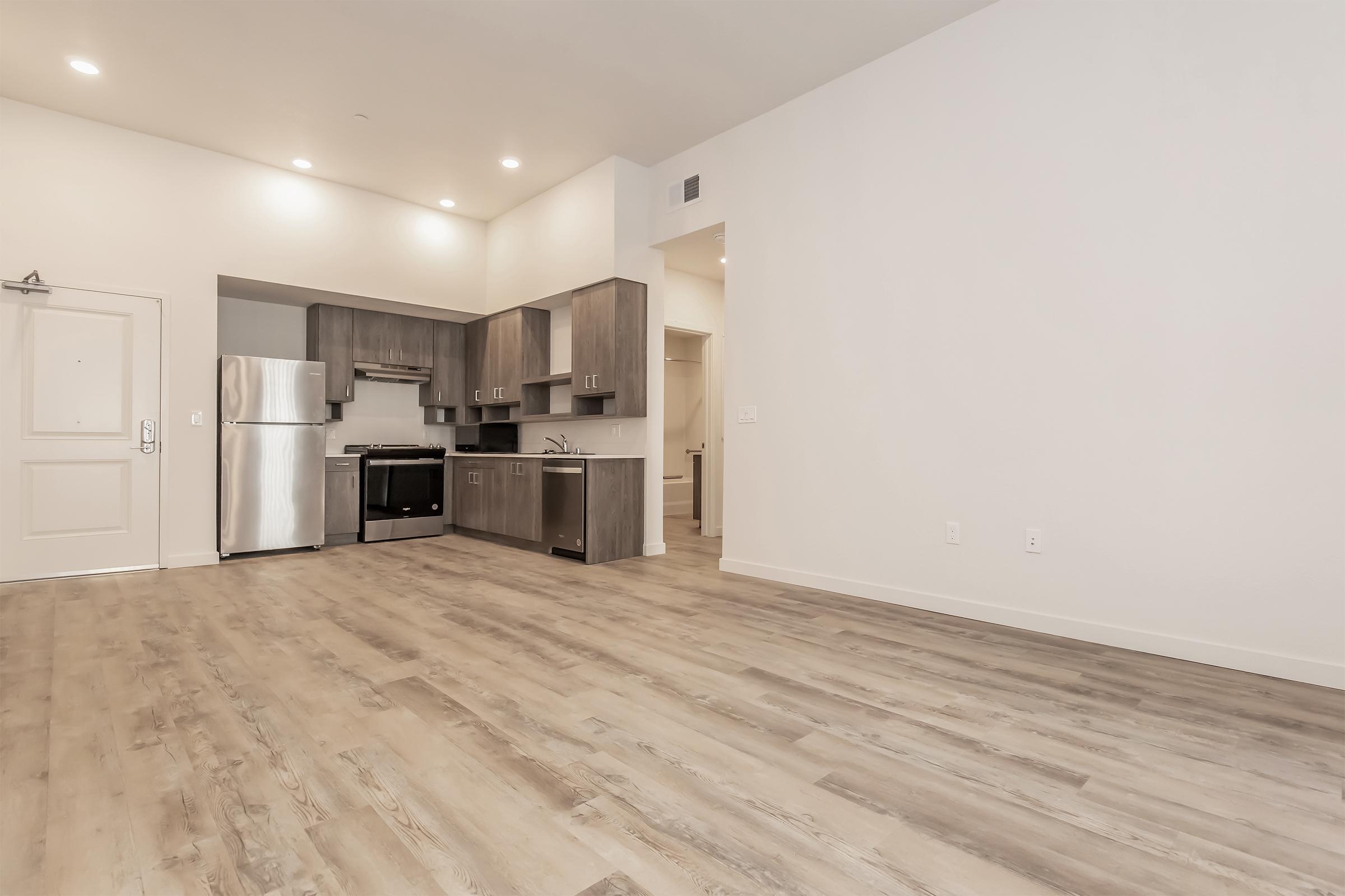 An open-concept modern kitchen and living area featuring light-colored wooden flooring, stainless steel appliances, and sleek cabinets. The space is well-lit with recessed lighting and a neutral color palette, creating a spacious and inviting atmosphere. A doorway leads to another room.