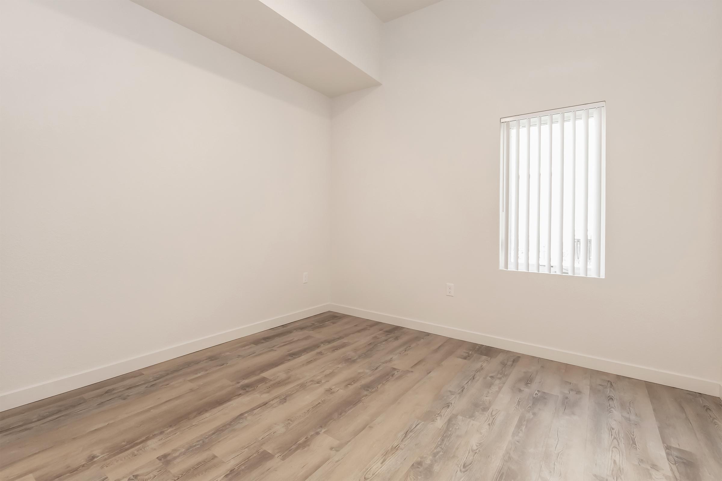 Empty room with light-colored walls and wooden flooring. A small window with vertical blinds allows natural light to enter. The space has a minimalist design, creating a clean and airy atmosphere.