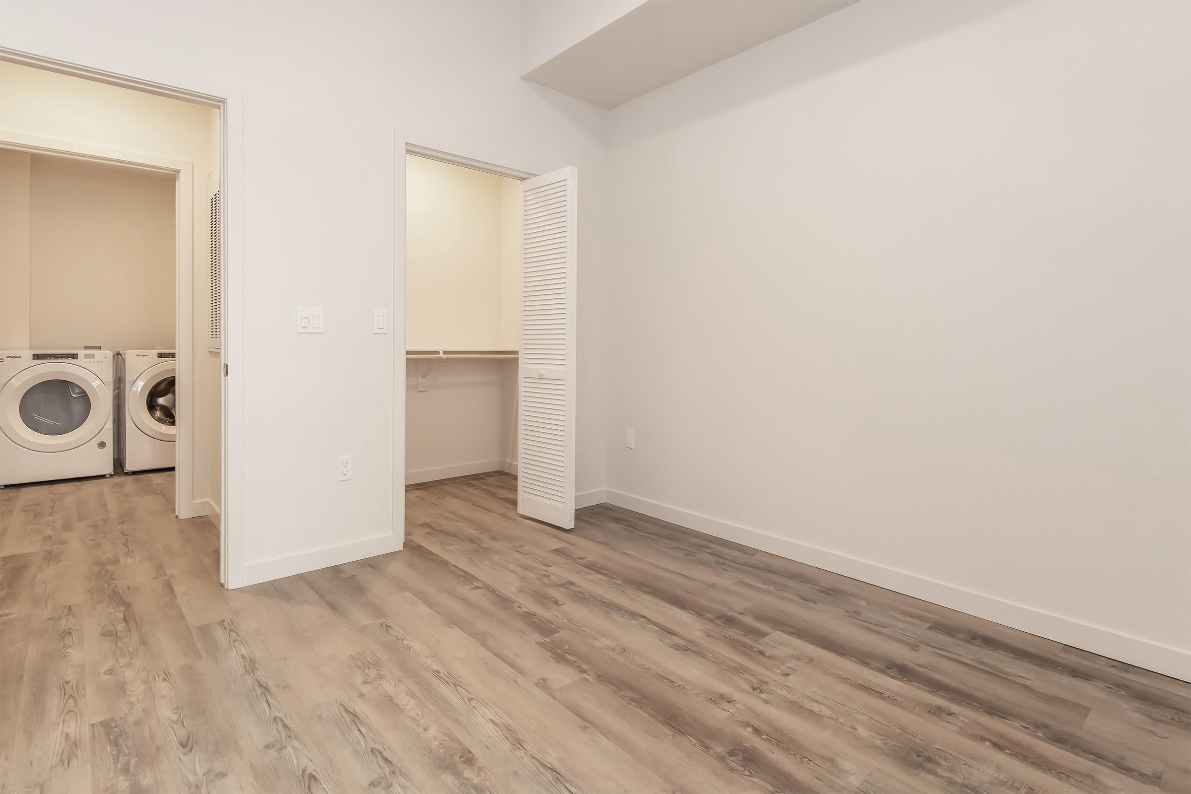 A spacious room featuring light wood flooring and a minimalistic design. On the left, there is a laundry area with stacked washer and dryer units. The room has two doors leading to other spaces, one of which is a closet. The walls are painted a soft neutral color, adding to the airy feel.