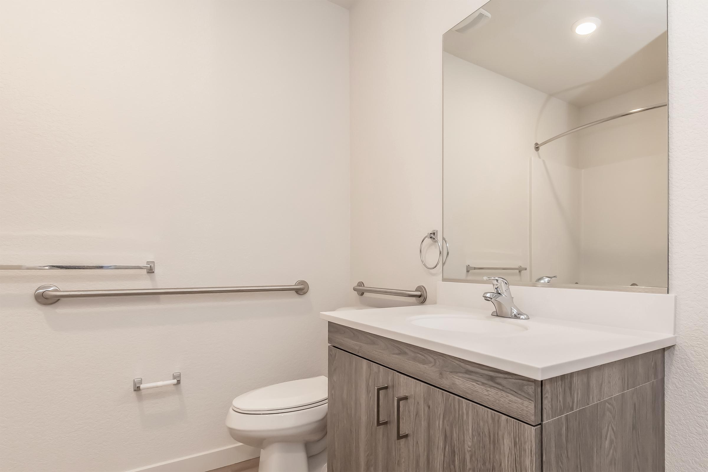 A clean, modern bathroom featuring a white toilet, a gray wooden cabinet with a sink, a wall mirror, and a shower area with a glass door. The walls are painted white, and there's a grab bar for accessibility. Bright overhead lighting illuminates the space.