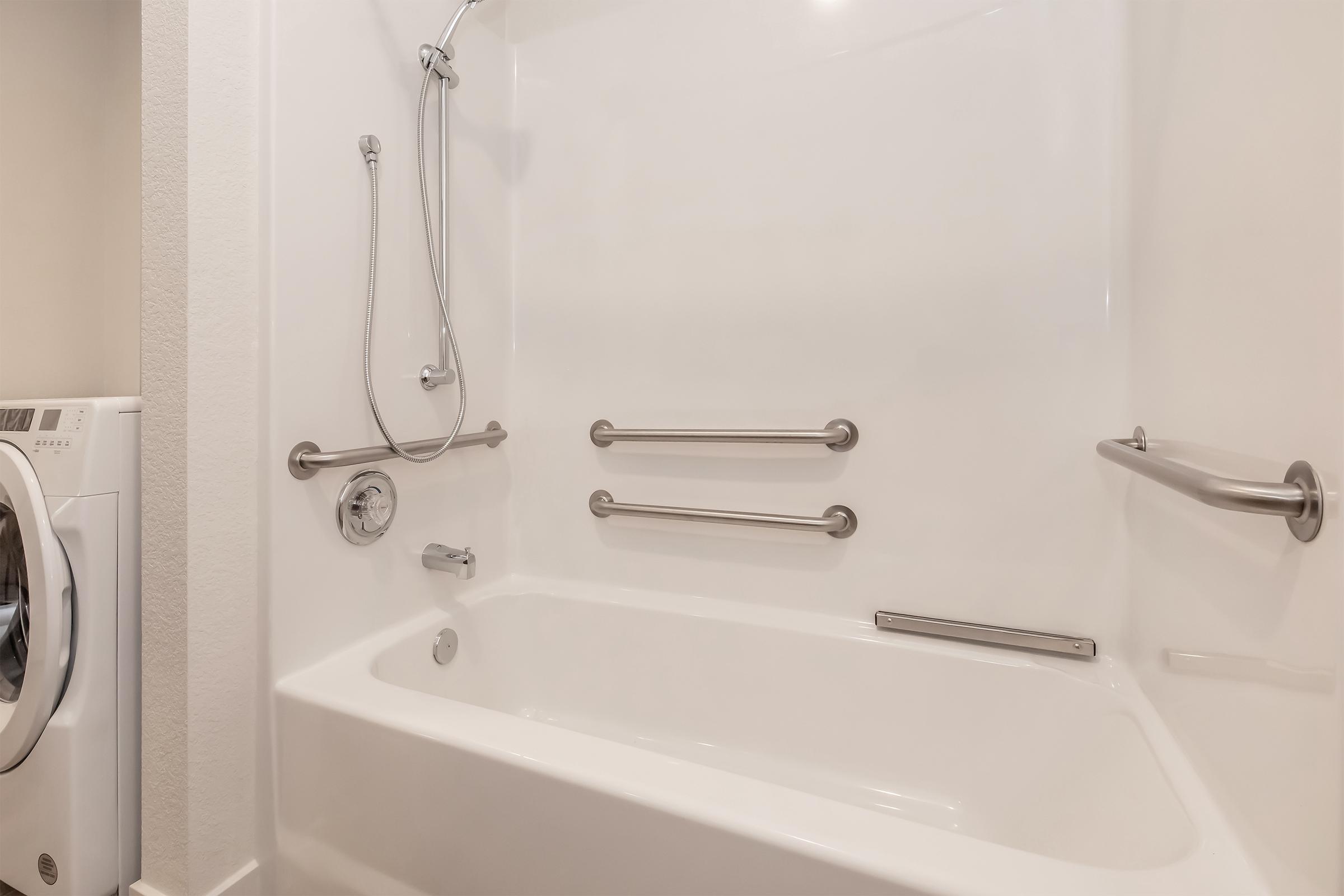 A clean and modern bathroom featuring a white bathtub with a built-in showerhead and grab bars for safety. A washing machine is visible to the left, blending with the contemporary design of the space. The walls are light-colored, contributing to a bright and open atmosphere.