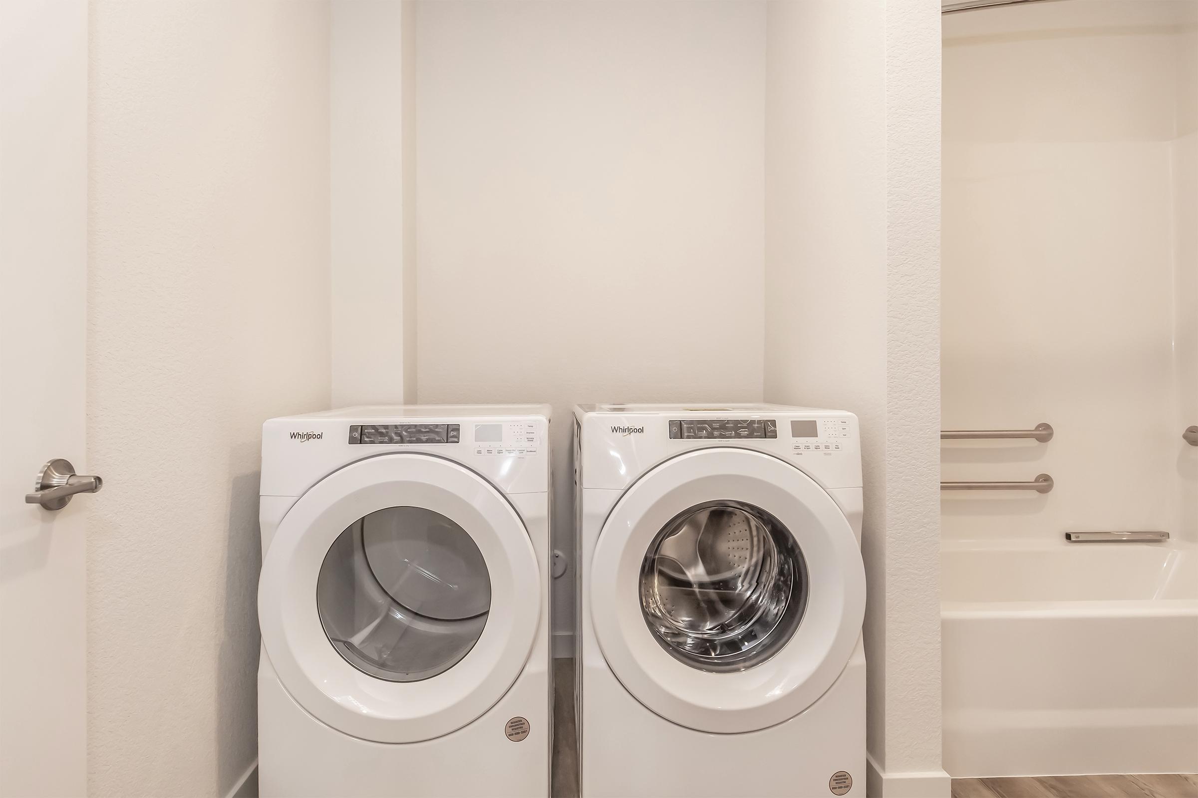 A bright laundry area featuring a stacked washer and dryer in white, positioned against a light-colored wall. Next to them is a bathtub with grab bars. The space is clean and modern, with light wood flooring and minimal decor.