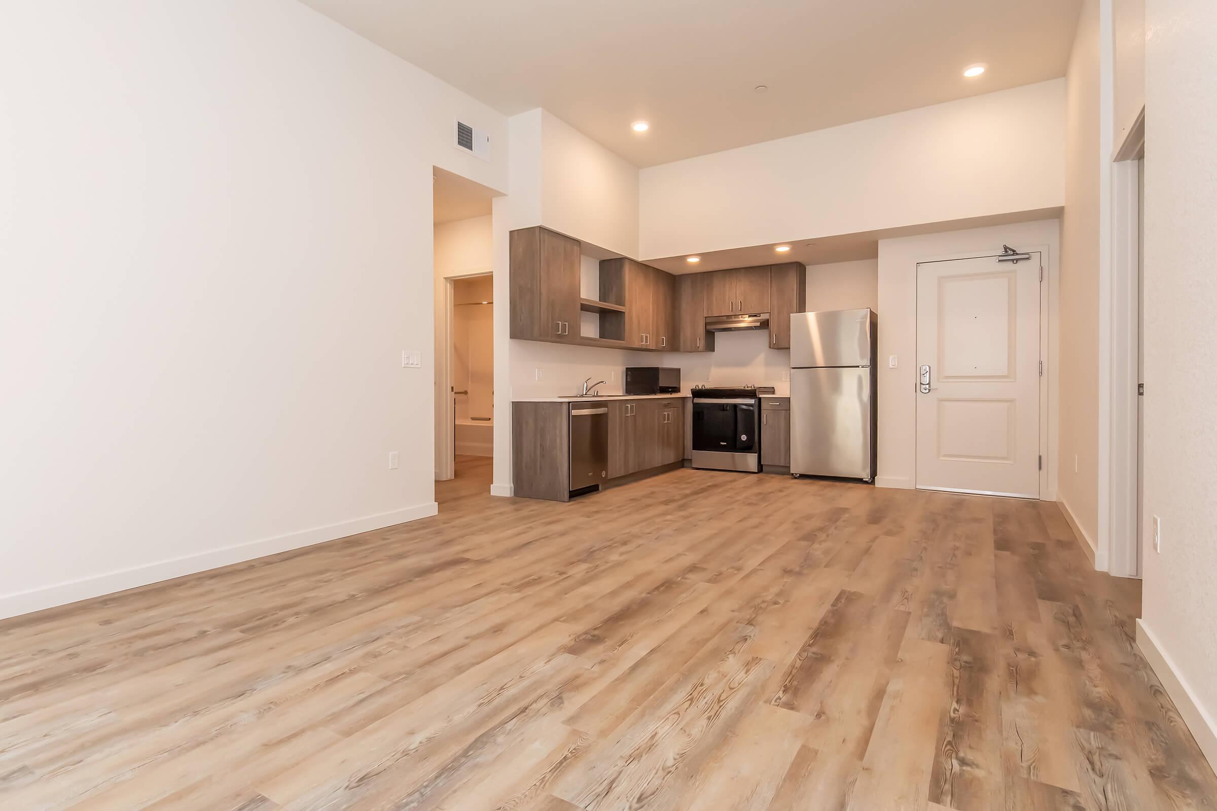 A modern kitchen and living area with an open layout. Features include wood-style flooring, stainless steel appliances, and wooden cabinets. The space is bright with neutral-colored walls and ample natural light coming from unseen windows, creating a spacious and inviting atmosphere.