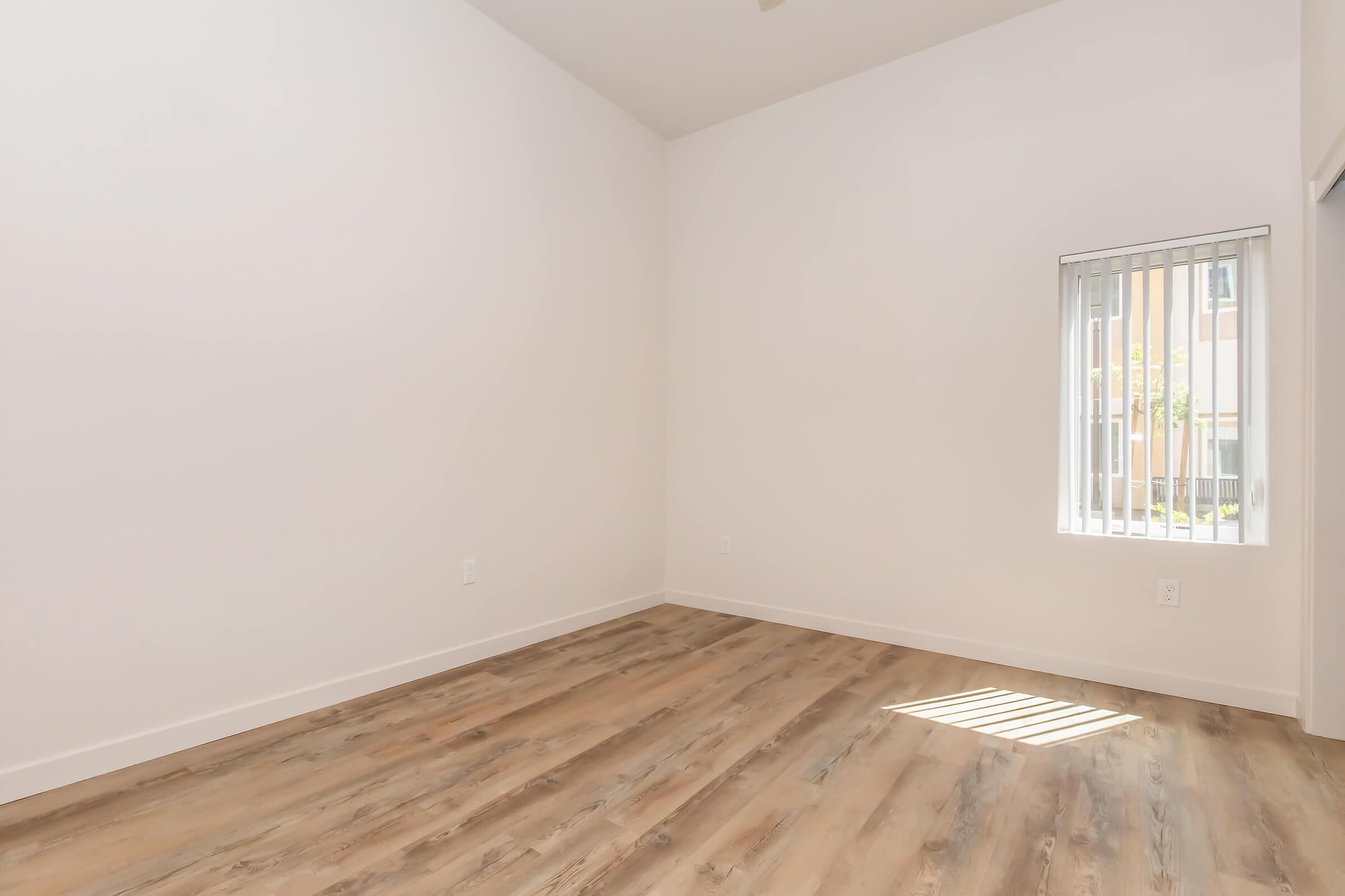 A vacant room with light wooden flooring, white walls, and a single window with vertical blinds letting in natural light. The room has high ceilings and minimal furnishings, creating an open and airy atmosphere.