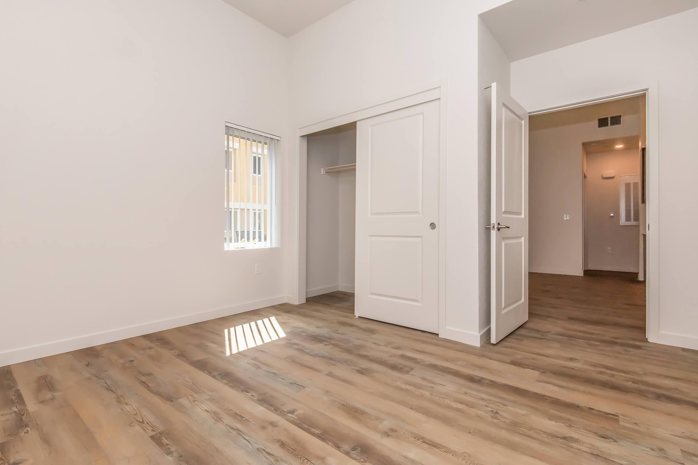 A spacious, empty room featuring light-colored walls and a large window with blinds. The room has a closet with sliding doors and a doorway leading to another area. The floor has a warm wood-like finish, creating a bright and airy feel. Natural light streams through the window, enhancing the open atmosphere.