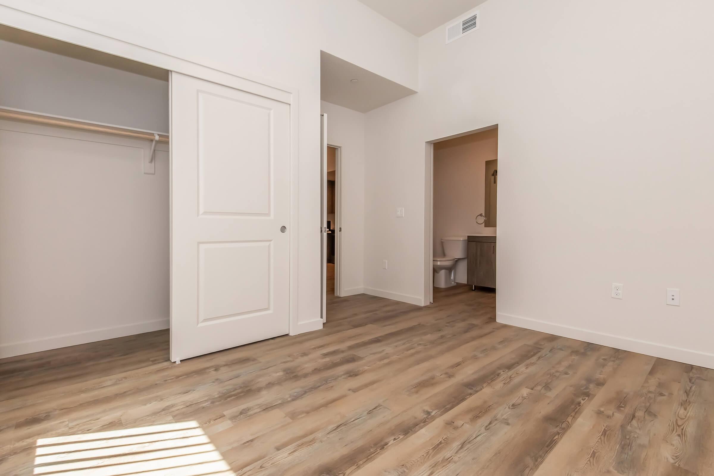 A spacious, empty room with light-colored walls and wooden flooring. There is a sliding closet door on the left and an open doorway leading to a bathroom on the right. The room is well-lit with natural light coming in, creating a bright and airy atmosphere.