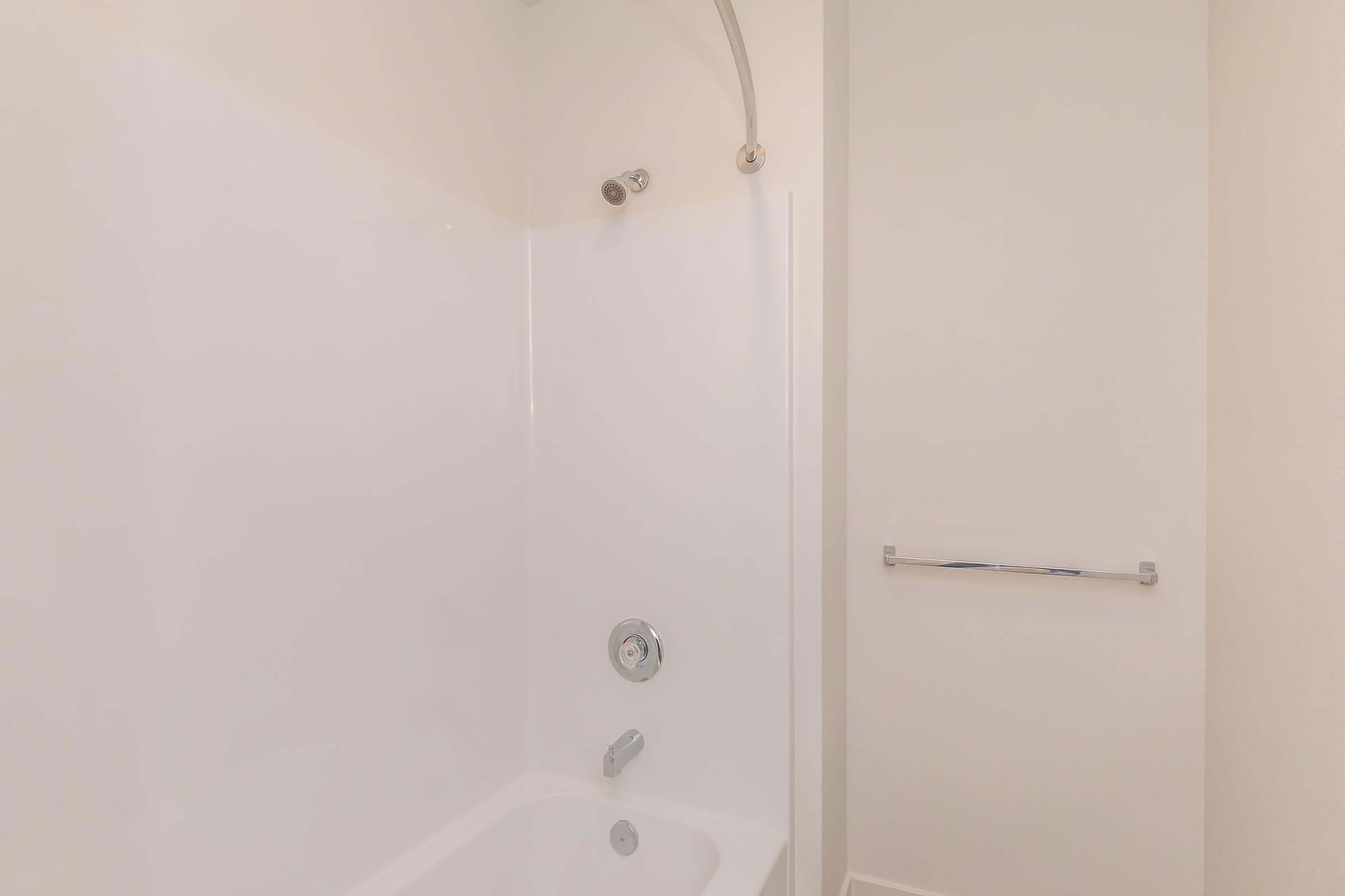 Interior view of a clean, modern bathroom showing a white shower stall with a chrome showerhead, a faucet, and a towel bar mounted on the wall. The walls are plain and light-colored, contributing to a minimalist and spacious aesthetic.