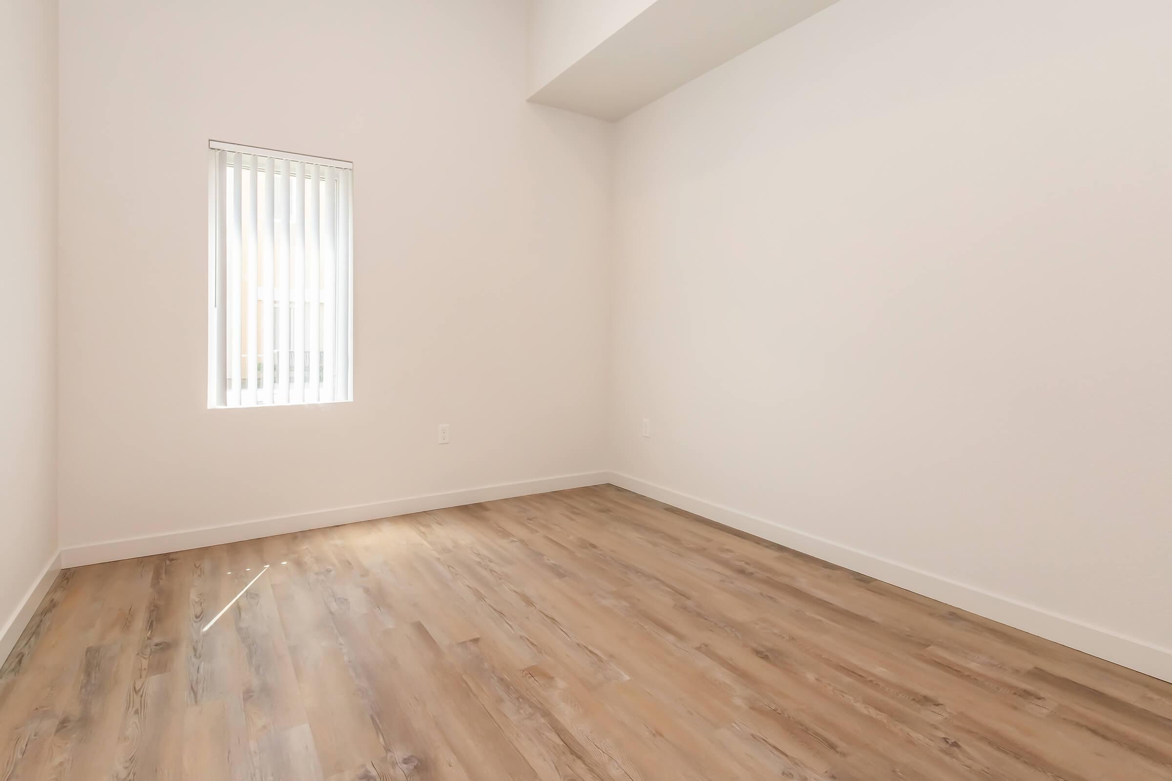 Empty room with white walls and a wooden floor. A small window with vertical blinds allows natural light to enter. The space is minimalistic and has a modern feel, ideal for various uses.
