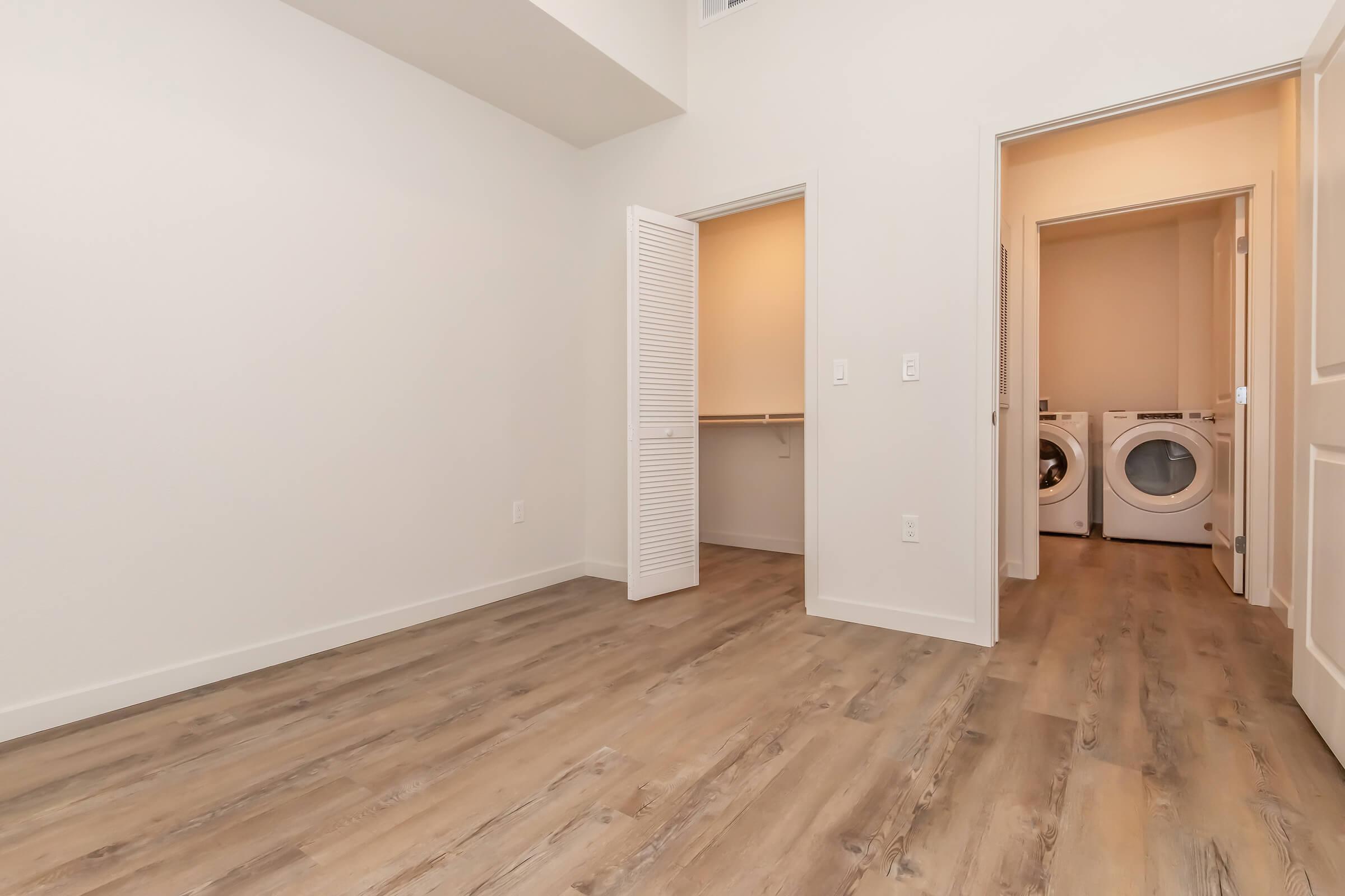 A spacious, well-lit room featuring light-colored walls and wooden flooring. There are two open doorways leading to adjacent spaces, one of which includes a laundry area with a washer and dryer. The room is empty, creating a minimalist aesthetic.