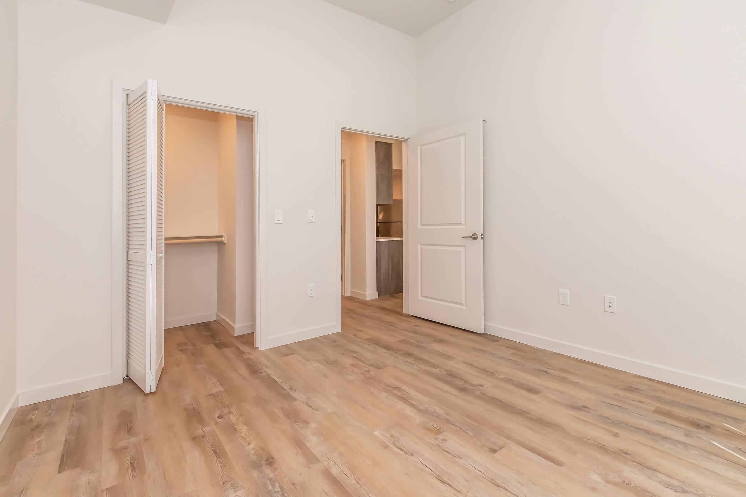 A bright and spacious room featuring light-colored walls and wooden flooring. There are two doorways: one leading to a closet and another to an adjacent space. The room appears well-lit, with natural light illuminating the interior.