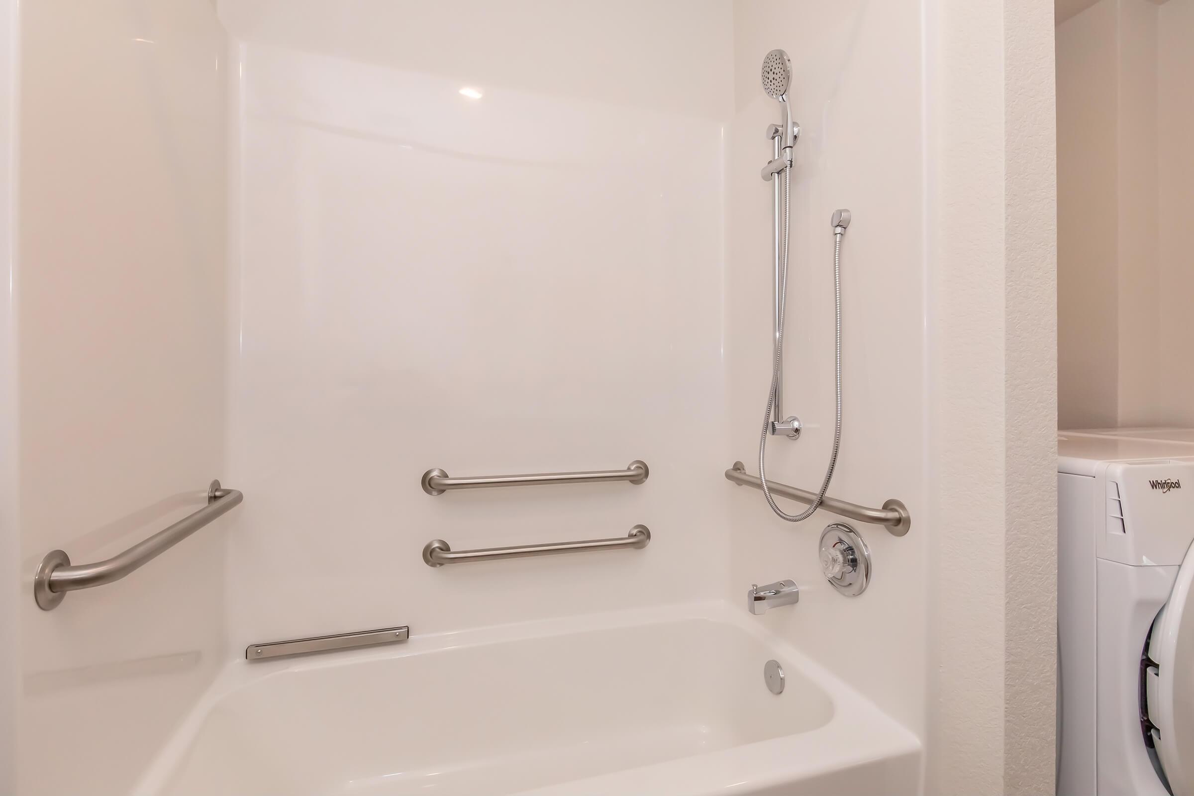A clean, white bathroom with a shower and bathtub combination. The shower has a handheld showerhead mounted on the wall, and there are safety grab bars installed along the walls. A washing machine is partially visible next to the tub, indicating a laundry area.