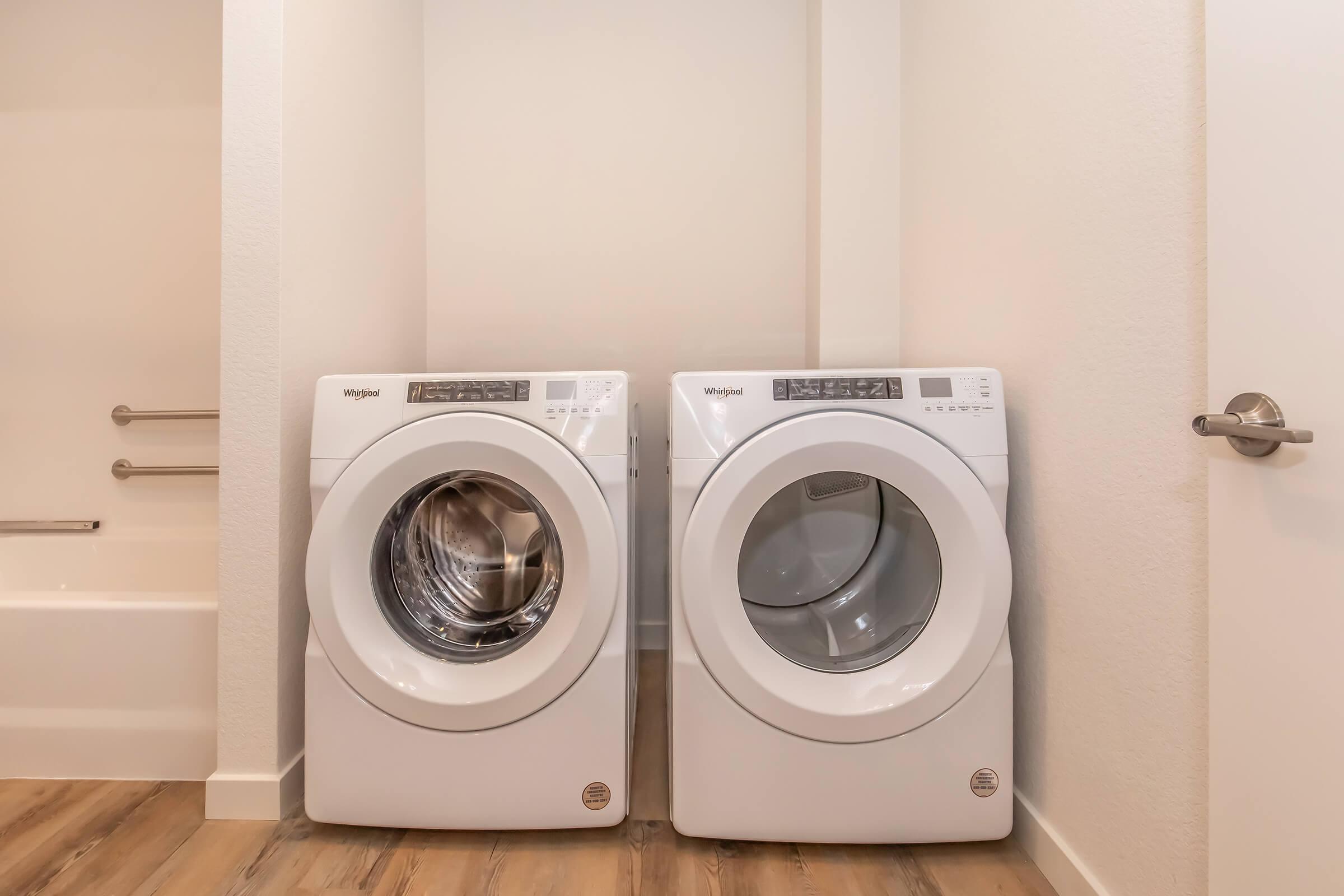 A laundry room featuring a stackable white washer and dryer set side by side on a wooden floor, with a light-colored wall and a door visible in the background. The appliances have digital control panels and a modern design, creating a clean and functional laundry space.