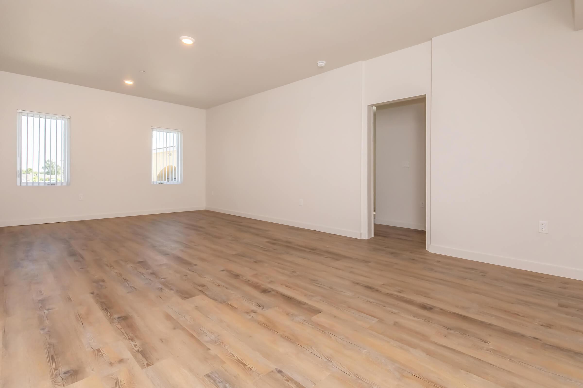 A bright, empty room with light-colored walls and large windows featuring horizontal blinds. The floor is made of laminate wood, giving a warm appearance. A doorway leads to another room, suggesting the space is part of a larger layout. The overall ambiance is open and airy.