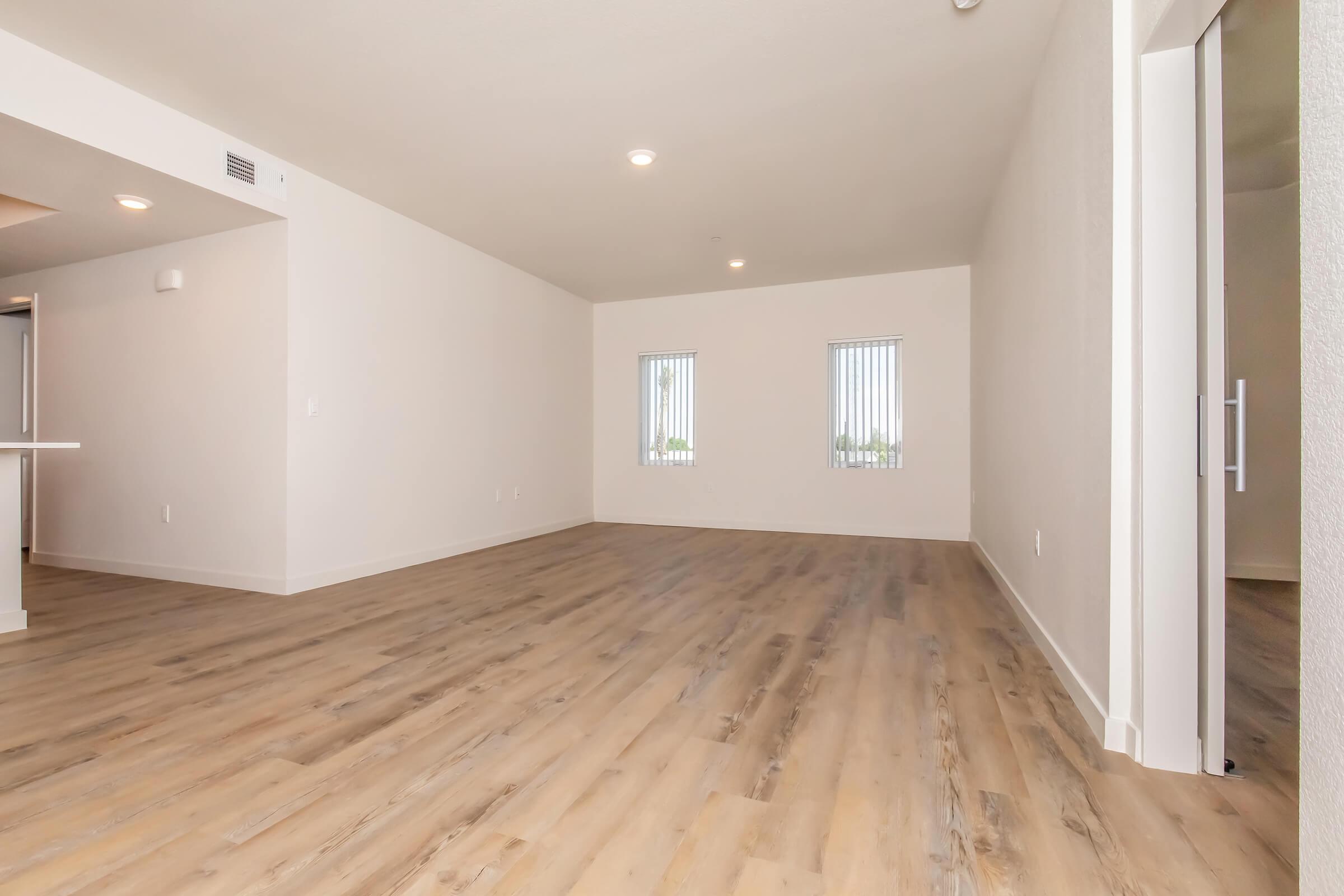 A spacious, empty room with light-colored walls and a wooden floor. Two vertical blinds-covered windows allow natural light to enter, and there is an open space leading to a kitchen area. The overall atmosphere is bright and inviting, suitable for residential living.