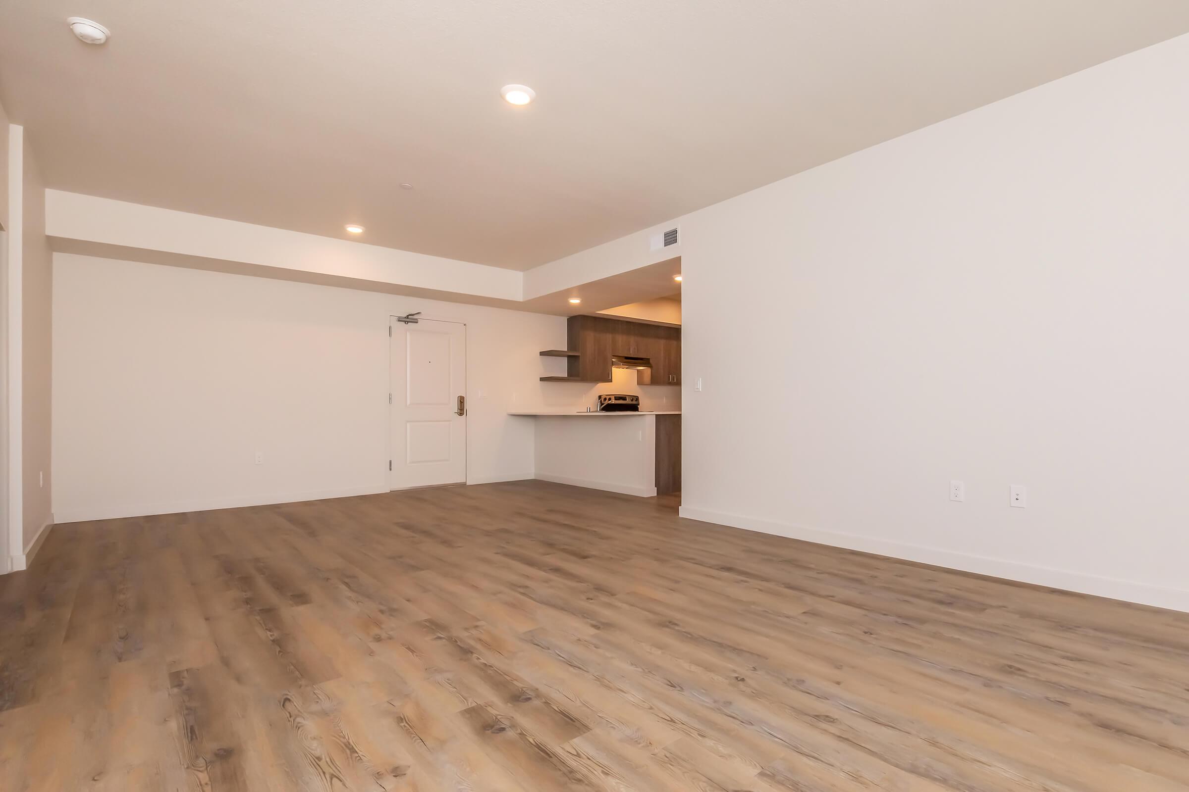 A spacious, empty room with light-colored walls and wood-like flooring. The room features a small kitchen area in the background, with modern cabinetry and countertops. Soft lighting and a neutral color palette create a bright and inviting atmosphere.