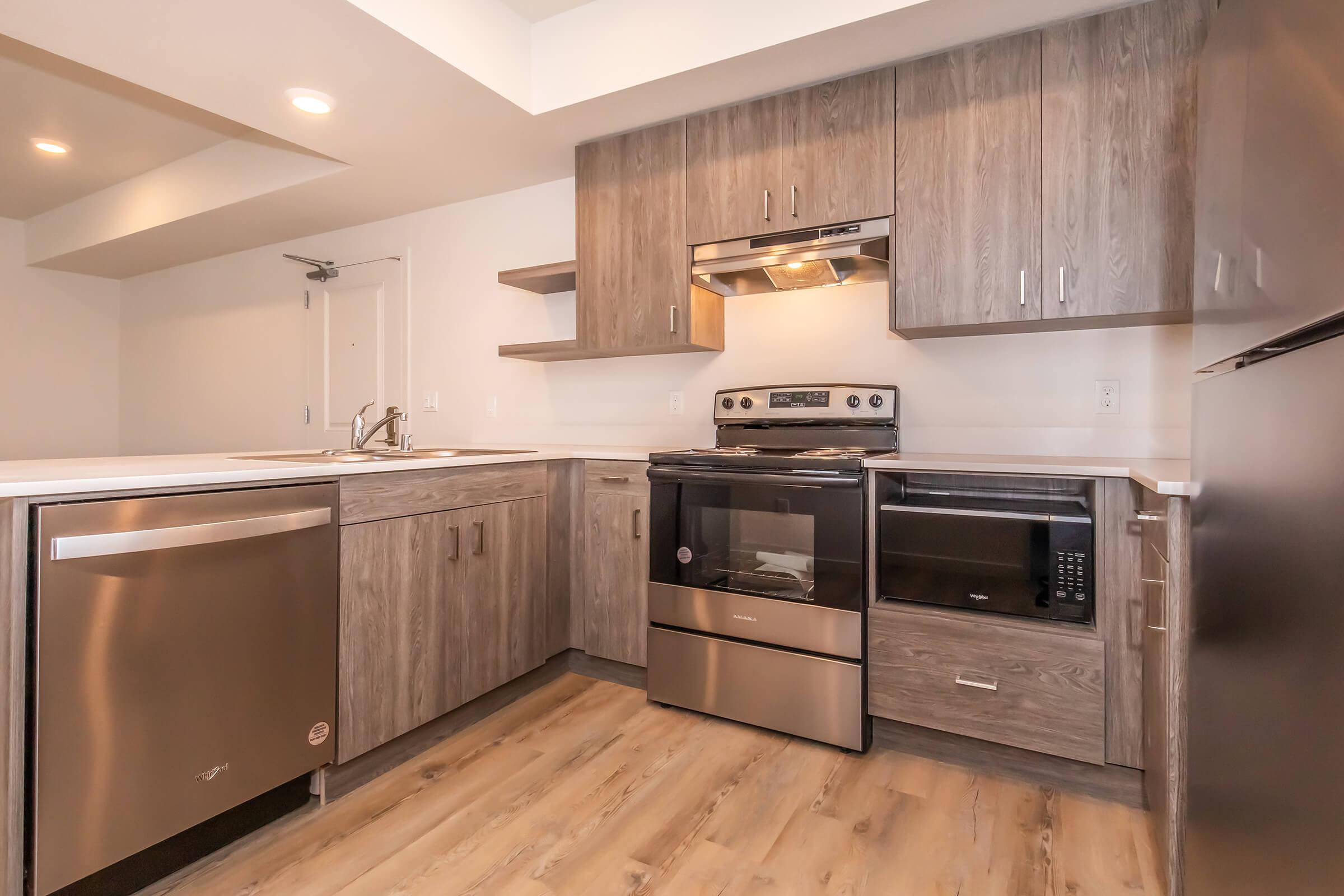 Modern kitchen featuring gray wooden cabinets, stainless steel appliances including a dishwasher, oven, and microwave, along with a sink and countertop space. The flooring is light-colored wood, and the overall design is sleek and contemporary with good lighting.