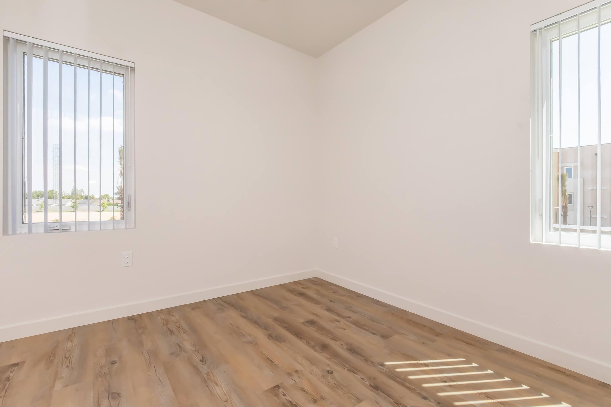 Empty room with light-colored walls, two windows with vertical blinds on opposite sides, and wooden flooring. Natural light enters through the windows, highlighting the spaciousness of the area.