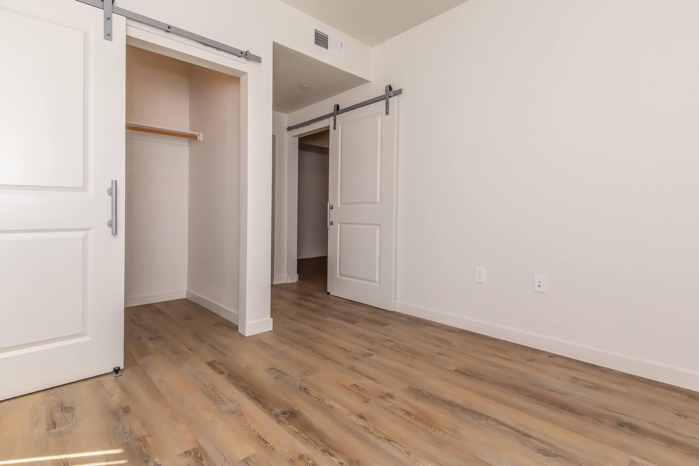 A spacious room with light-colored walls and a wood plank floor. There are two sliding doors: one leads to a closet with a shelf, and the other opens to an adjoining space. The room is well-lit and has a modern, clean aesthetic.