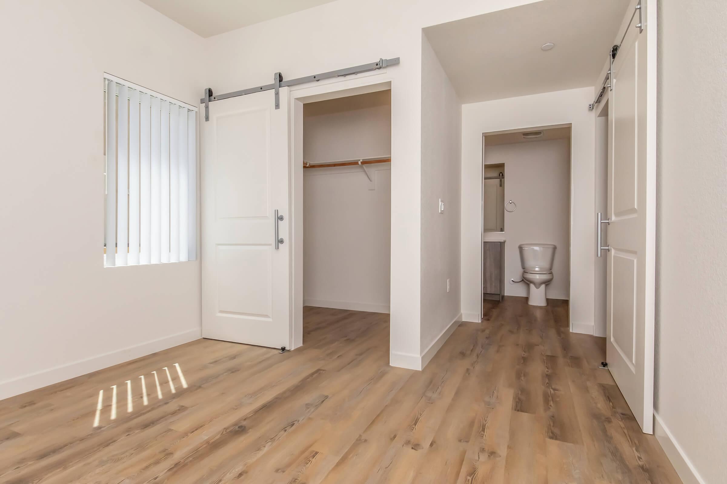 A spacious, modern room featuring a large window with blinds, wooden flooring, and white walls. To the left, there is a sliding door leading to a closet, while a hallway on the right leads to a bathroom with a toilet. The layout offers a bright and airy feel, suitable for various uses.