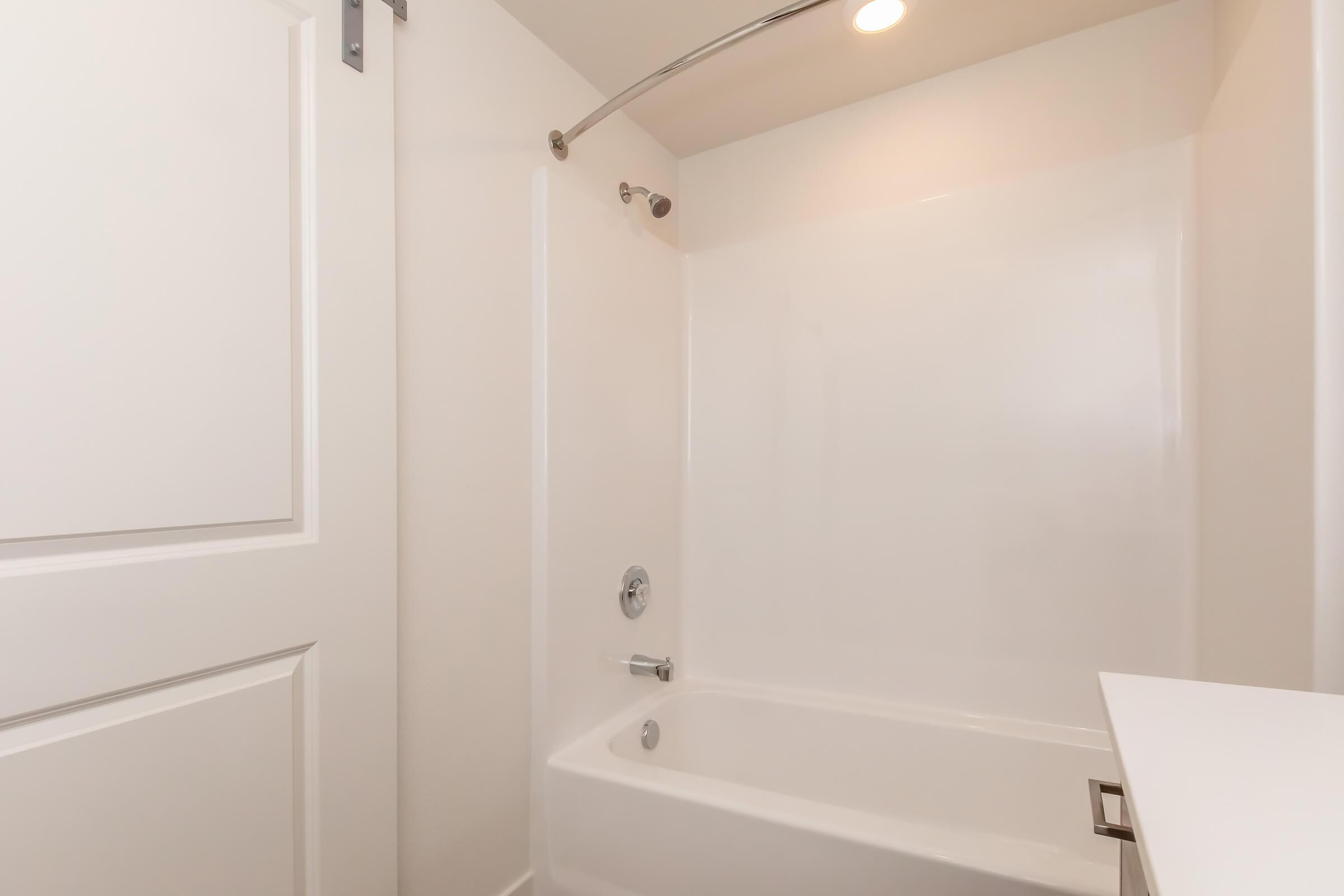 A clean and simple bathroom featuring a white bathtub and shower combination. The walls are smooth with a light finish, and there is a metal shower rod. A bright overhead light illuminates the space, contributing to the modern and minimalistic design. A closed white door can also be seen in the frame.