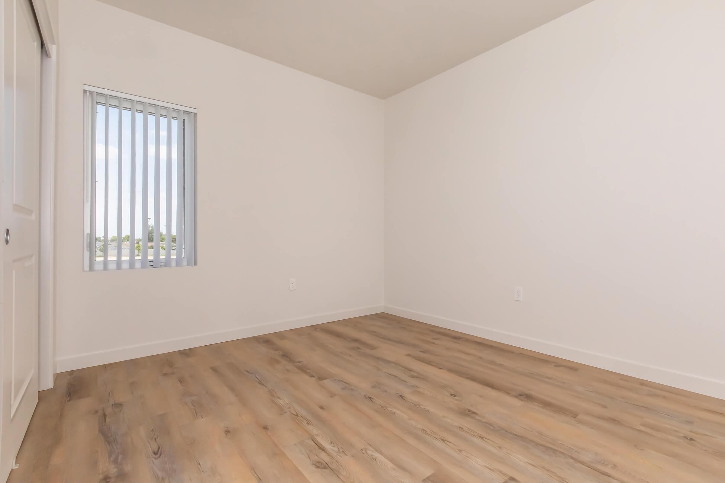 A spacious, empty room with light-colored walls and a light wood floor. It features a vertical blind window allowing natural light, and a closet with sliding doors. The space is unfurnished, providing a blank canvas for customization.