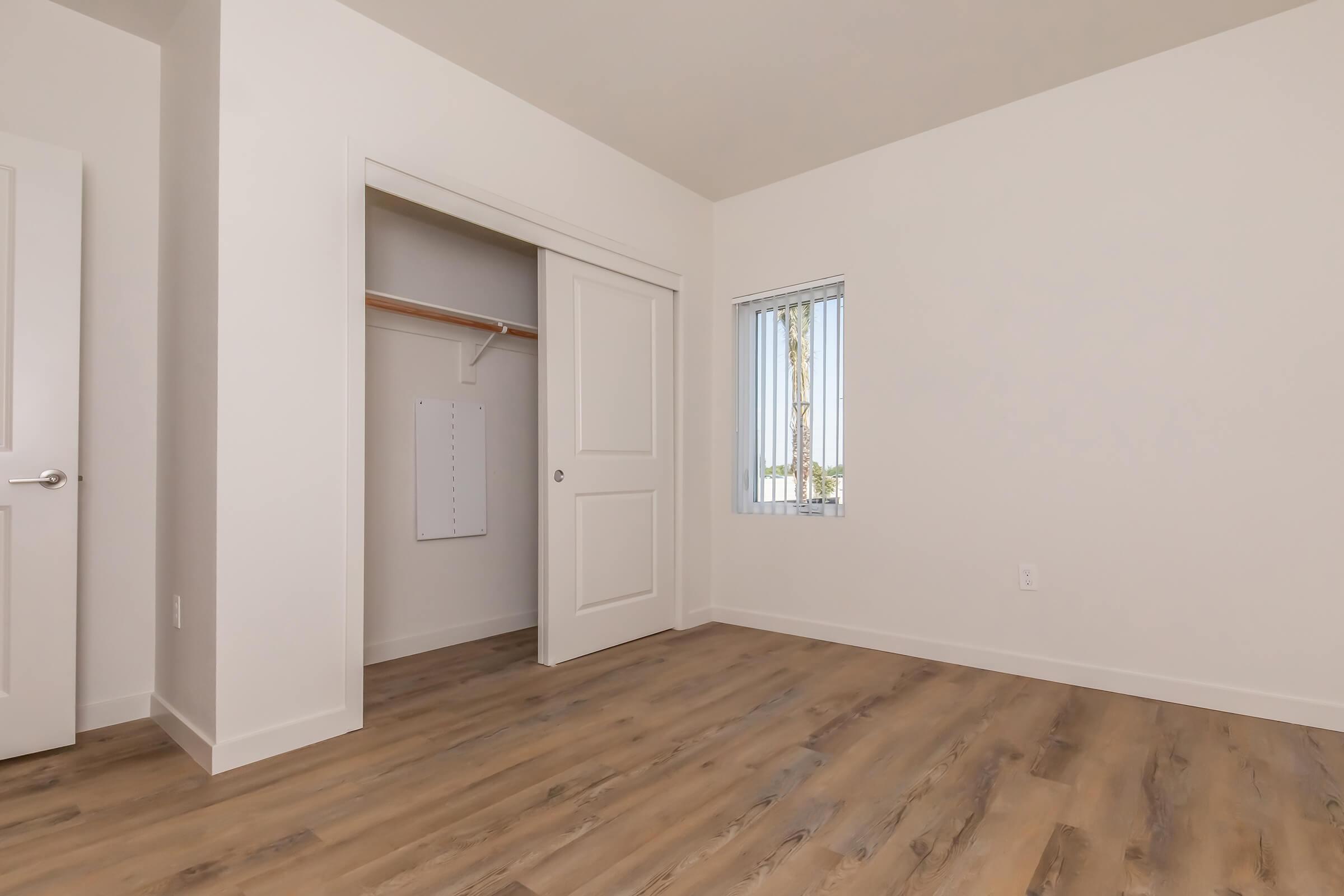 A minimalistic room featuring light-colored walls and a wooden floor. On the left, there is a closet with a sliding door, and to the right, a small window with vertical blinds allows natural light to enter. The space appears clean and empty, suitable for various interior design options.