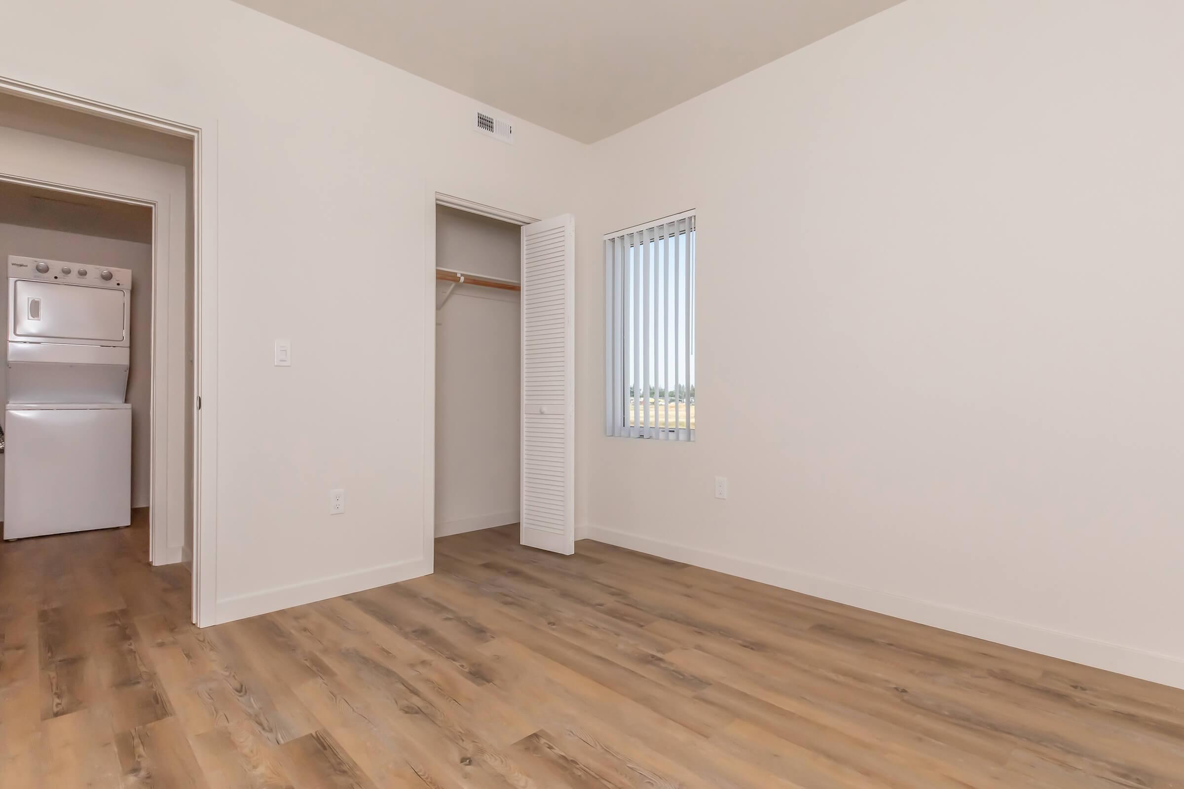 A bright, empty room with white walls and a wooden floor. A closet with folding doors is visible, as well as an open doorway leading to a laundry area with a stacked washer and dryer. Large windows with horizontal blinds allow natural light to enter the space.