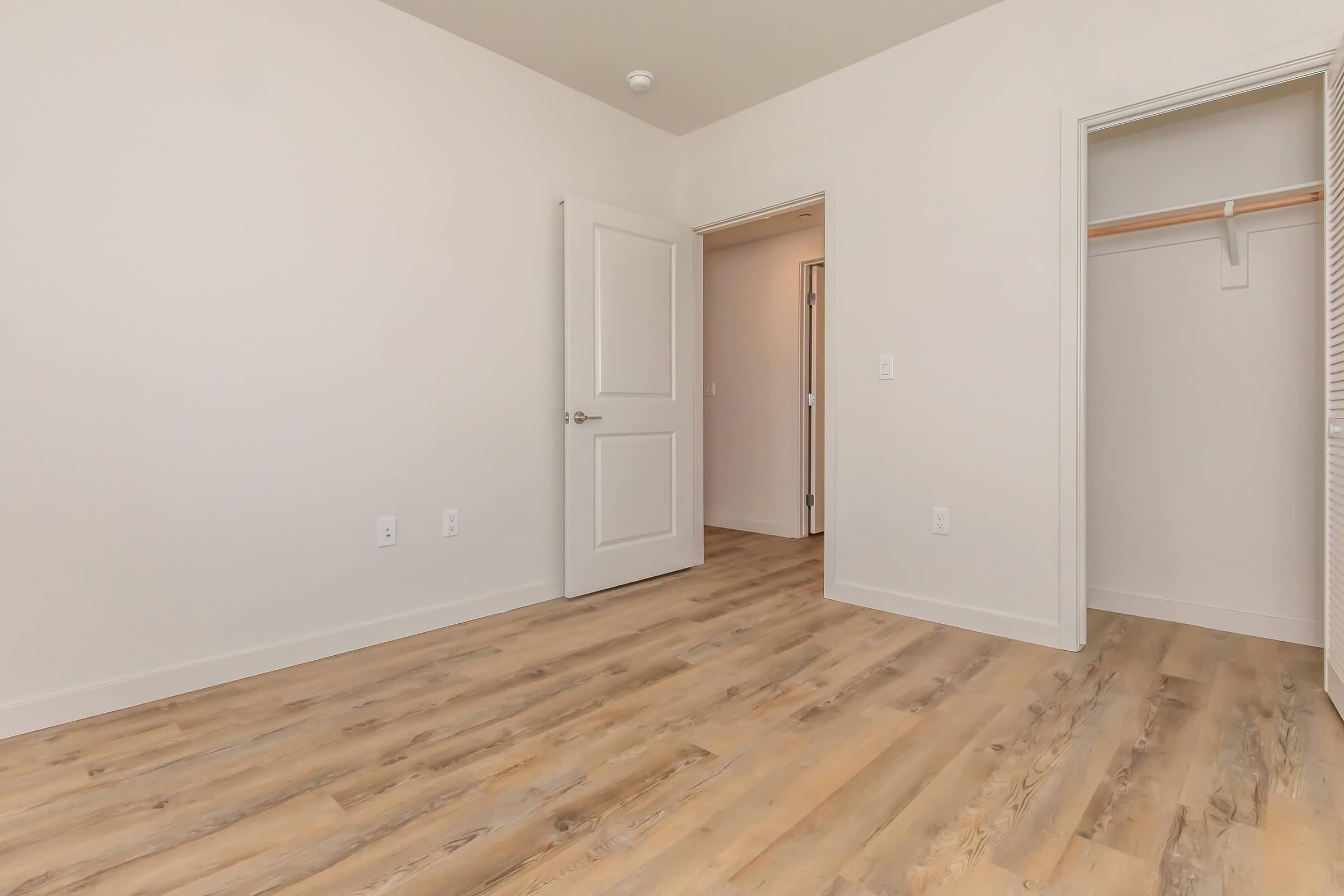 A spacious, empty room featuring light-colored walls, a wooden floor, an open door leading to a bathroom, and a closet with a single shelf. The room is well-lit and has no furniture, emphasizing a clean, minimalist appearance.
