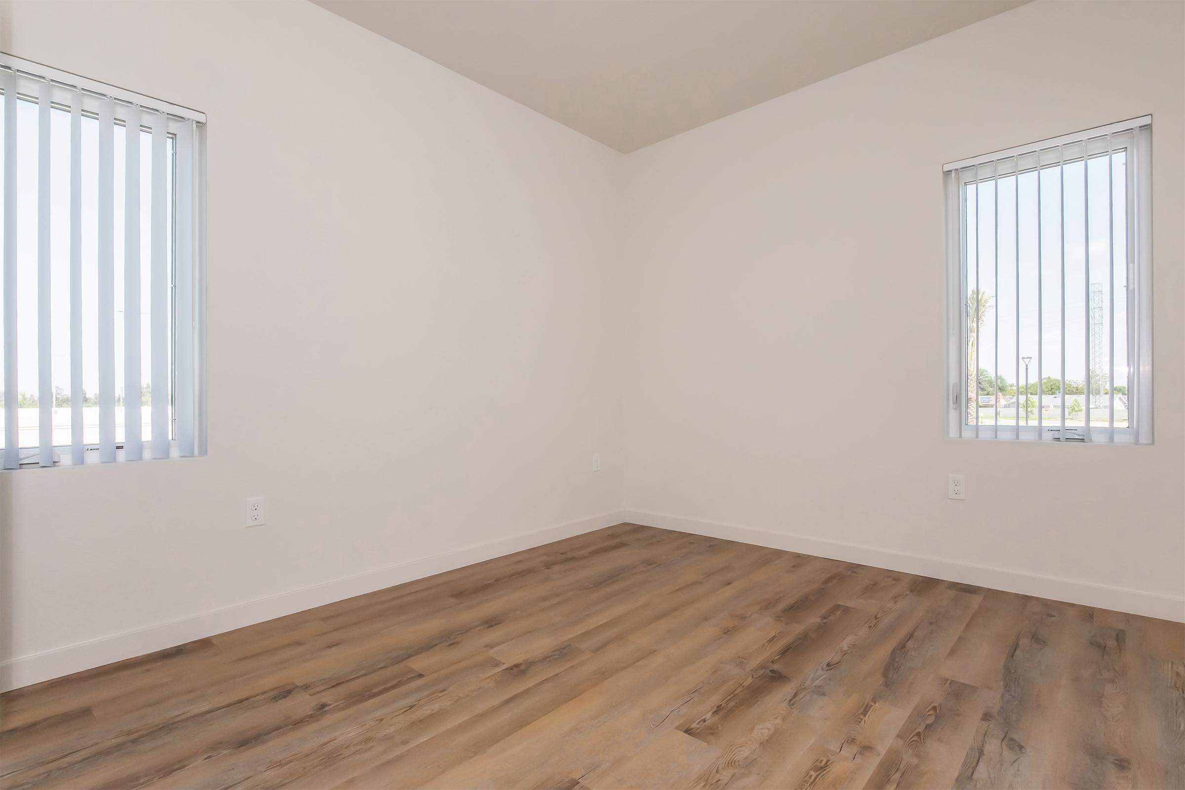 A minimalistic room featuring light-colored walls and wooden flooring. Two windows with vertical blinds allow natural light to enter the space, which is empty and spacious, suitable for various interior designs. The overall aesthetic is clean and modern.