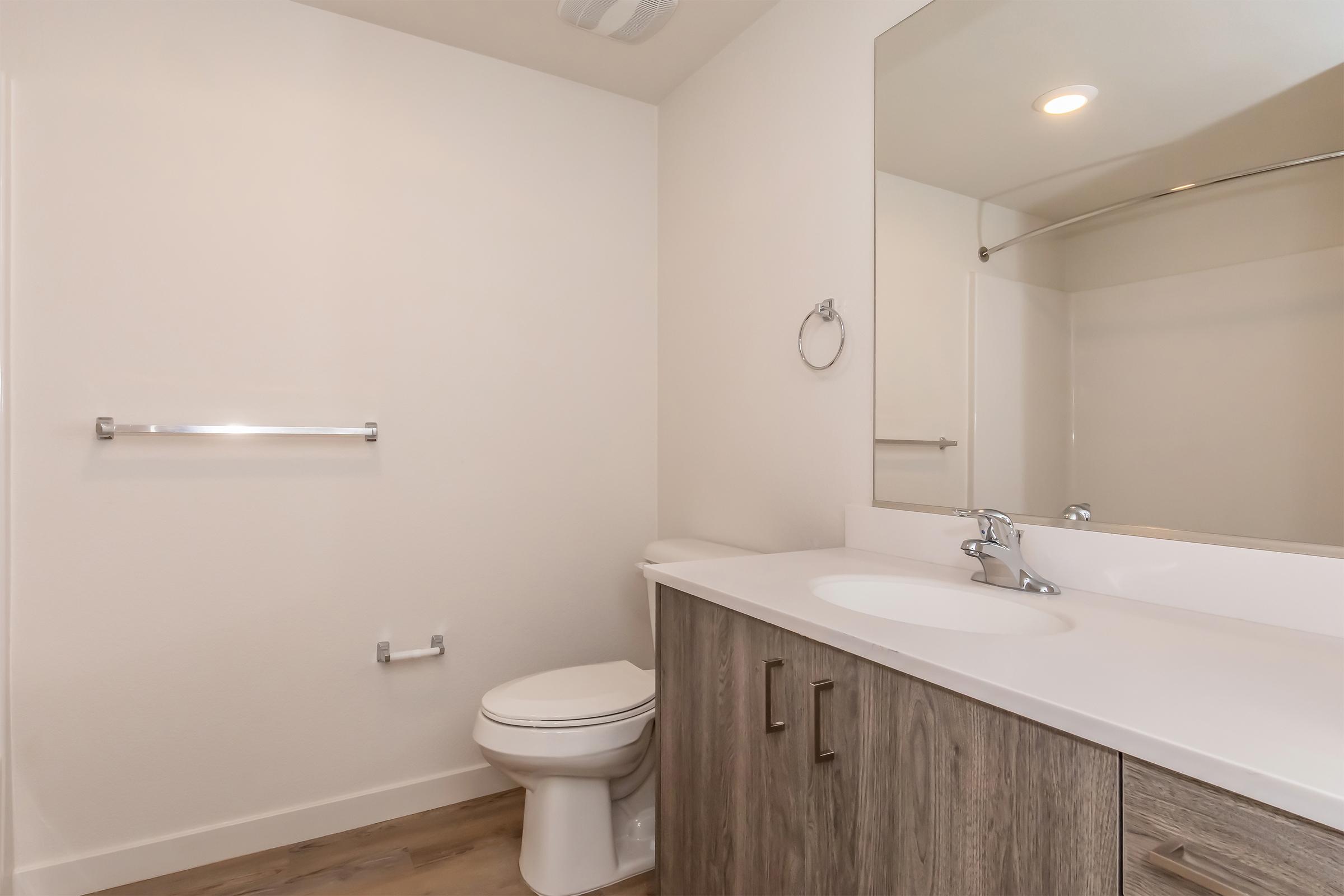Modern bathroom featuring a white toilet, a sleek vanity with a sink, and a large mirror above it. Neutral-colored walls and wood-like flooring create a clean and minimalist aesthetic. A towel rack is installed on the wall, and natural light complements the space.