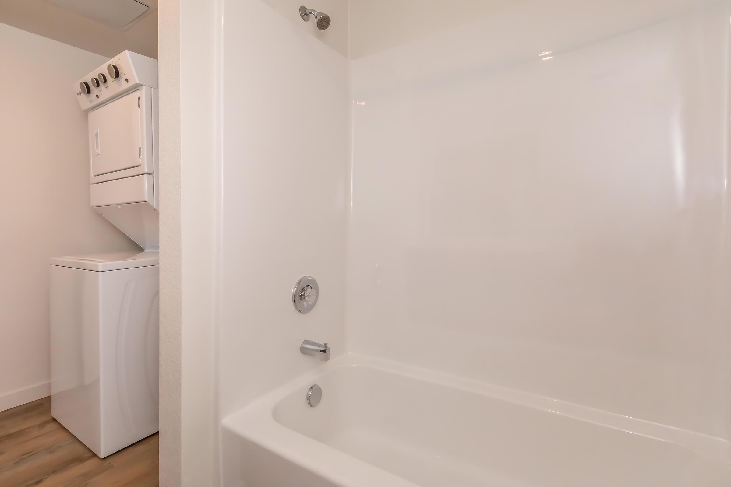 A clean, modern bathroom featuring a bathtub with a shower fixture. Next to the bathtub, there is a stacked washer and dryer appliance. The walls are white, and the floor has a wood-like finish, creating a bright and spacious atmosphere.
