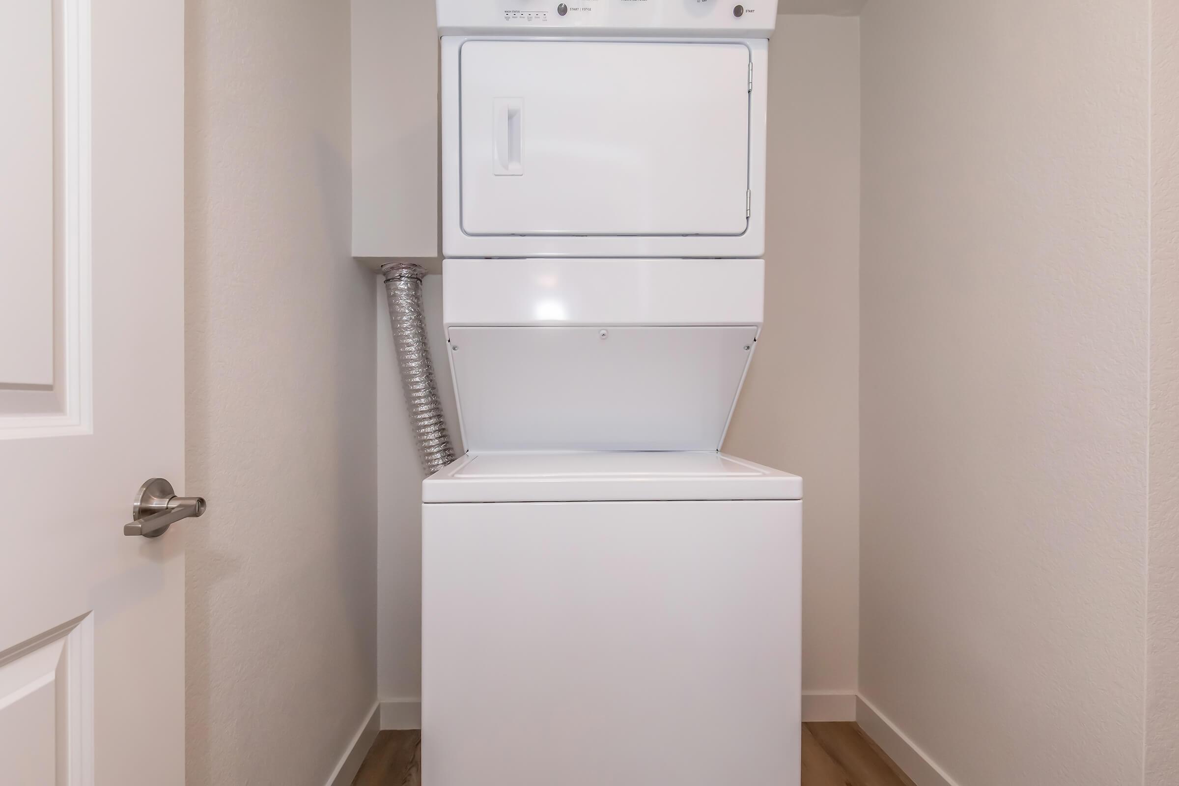 A stacked washer and dryer unit in a small laundry area, with a light-colored wall and wooden floor. The door to the laundry space is partially visible on the left.