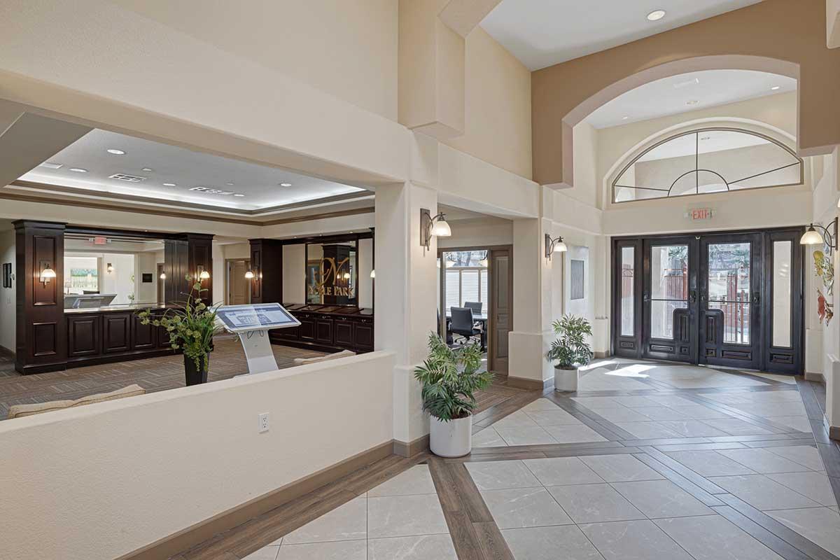 Interior of a modern lobby featuring a spacious design with a large entrance, elegant furnishings, potted plants, and a digital information panel. Bright lighting enhances the welcoming atmosphere, while dark wood accents add a touch of sophistication. Wide glass doors lead outside to a scenic view.
