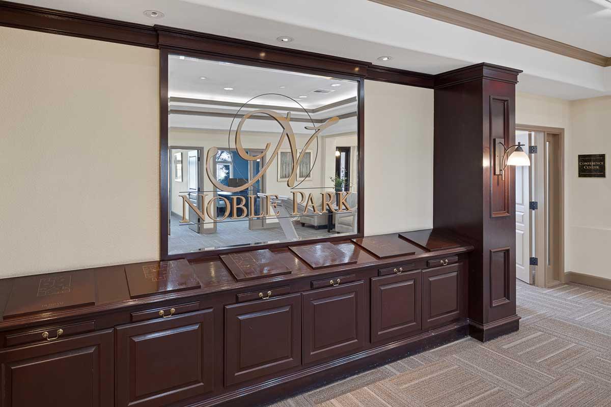 The image shows a stylish reception area with a polished wooden counter featuring engraved plaques. A large mirror with the logo "NOBLE PARK" is mounted on the wall, reflecting the elegant design of the space. Soft lighting enhances the warm colors and decor, creating a welcoming atmosphere.