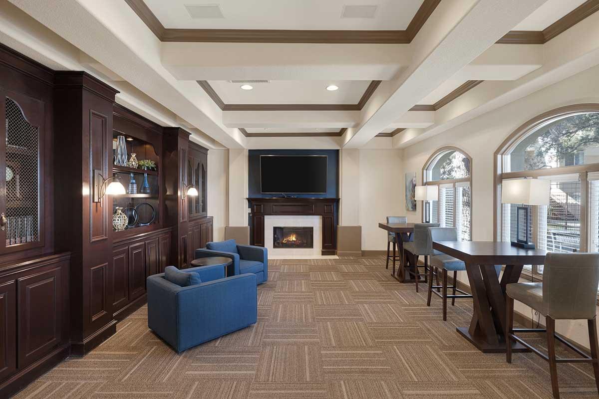 A cozy lounge area featuring a fireplace and a large flat-screen TV mounted on the wall. There are two blue armchairs and two tables with tall chairs, along with built-in shelving showcasing decorative items. Large windows allow natural light, enhancing the warm, inviting atmosphere of the space.