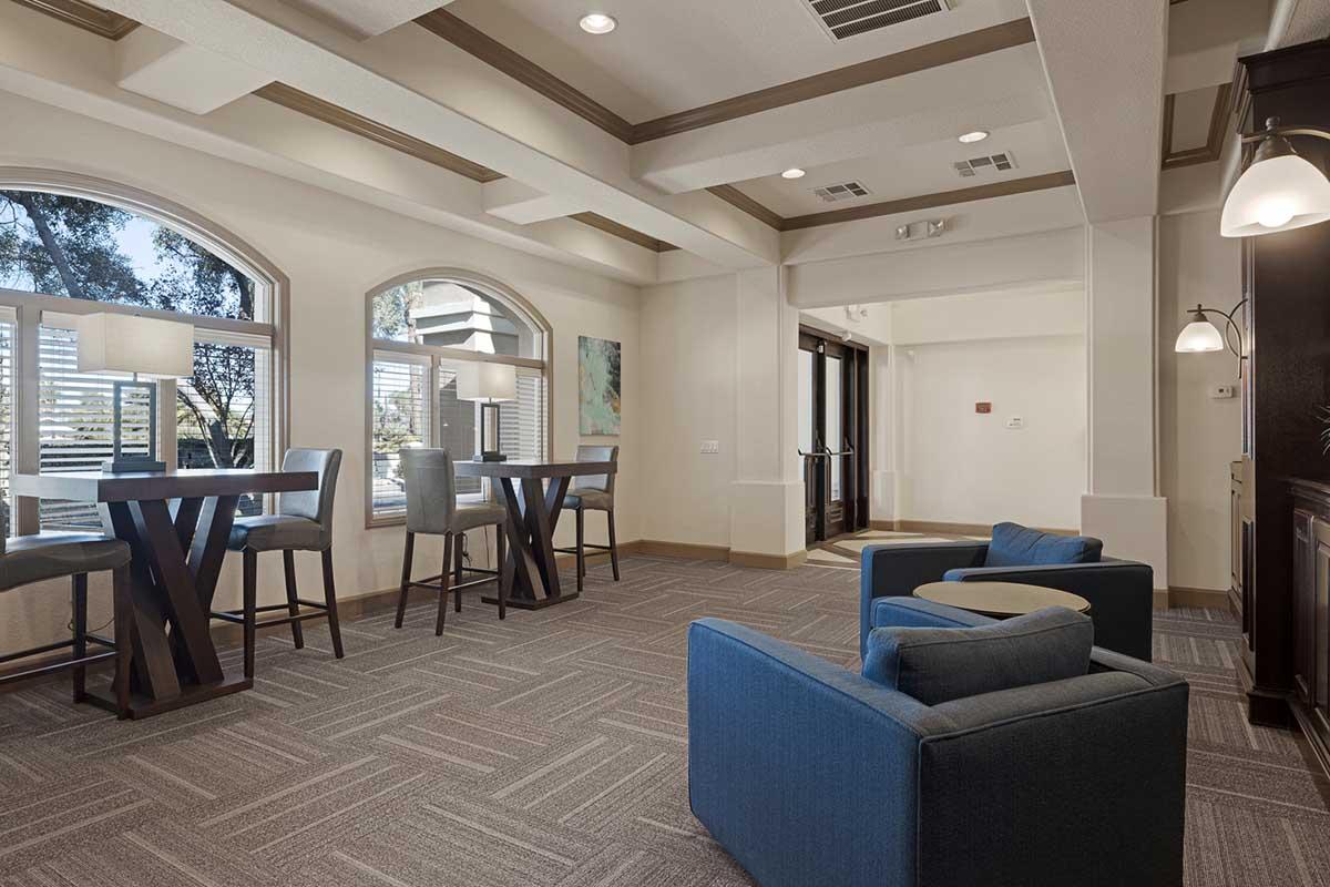 A modern lounge area featuring two blue chairs and high tables with stools, surrounded by large windows with natural light. The space has a neutral color palette, decorative ceiling details, and warm lighting fixtures, creating a welcoming and stylish atmosphere.