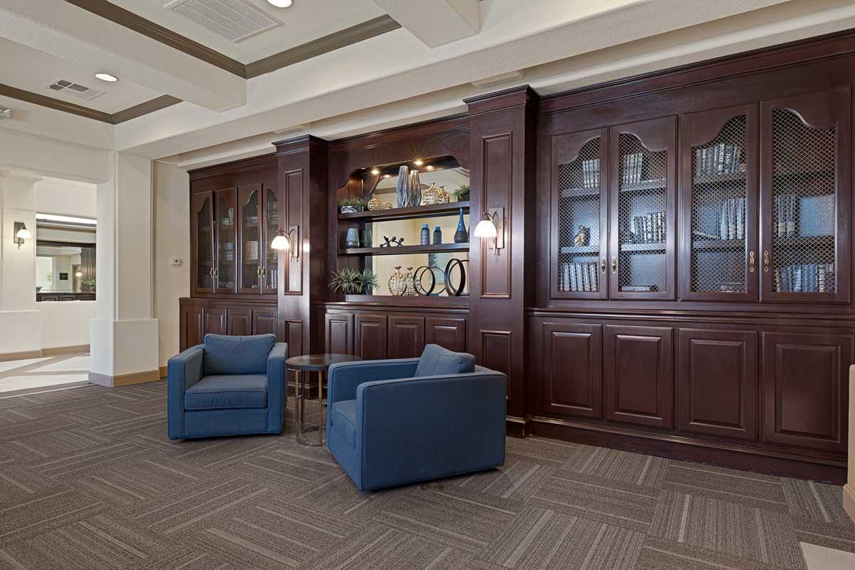 A cozy interior scene featuring two blue armchairs facing a wooden bookshelf with glass doors. The bookshelf is decorated with various items and plants, and there are wall sconces providing soft lighting. The flooring is a neutral carpet, and the overall atmosphere is inviting and warm.
