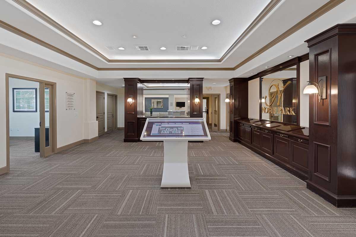 Interior view of a modern lobby featuring a digital directory on a pedestal. The space is well-lit with a stylish design, including dark wood accents and elegant lighting. A reception area is visible in the background, along with a logo on the wall. The flooring is a textured brown pattern.
