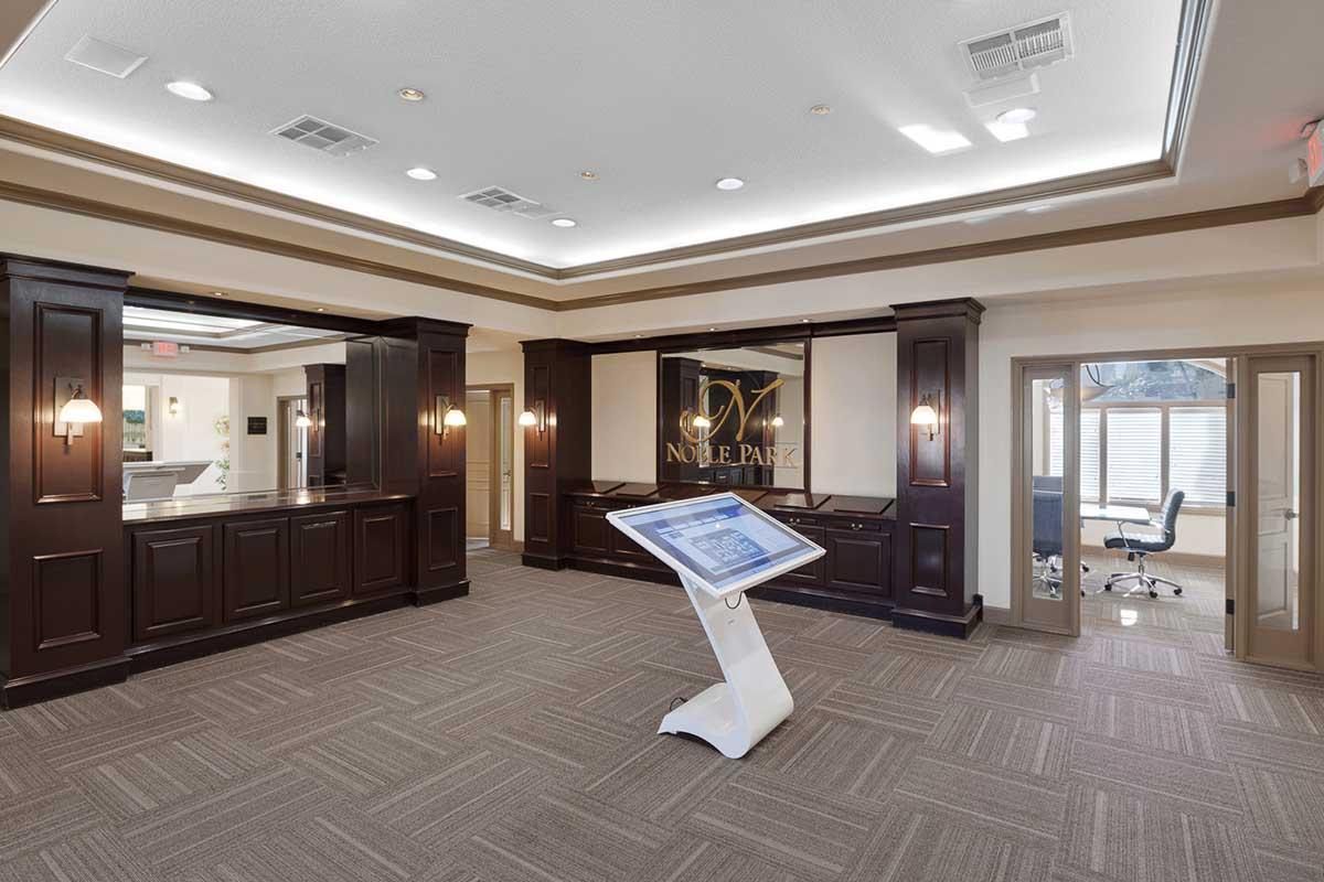 Interior view of a modern reception area featuring dark wooden cabinetry, a digital touchscreen kiosk, and spacious seating. Natural light floods in through large windows, contributing to a welcoming atmosphere. The design is contemporary, with neutral tones and soft lighting.
