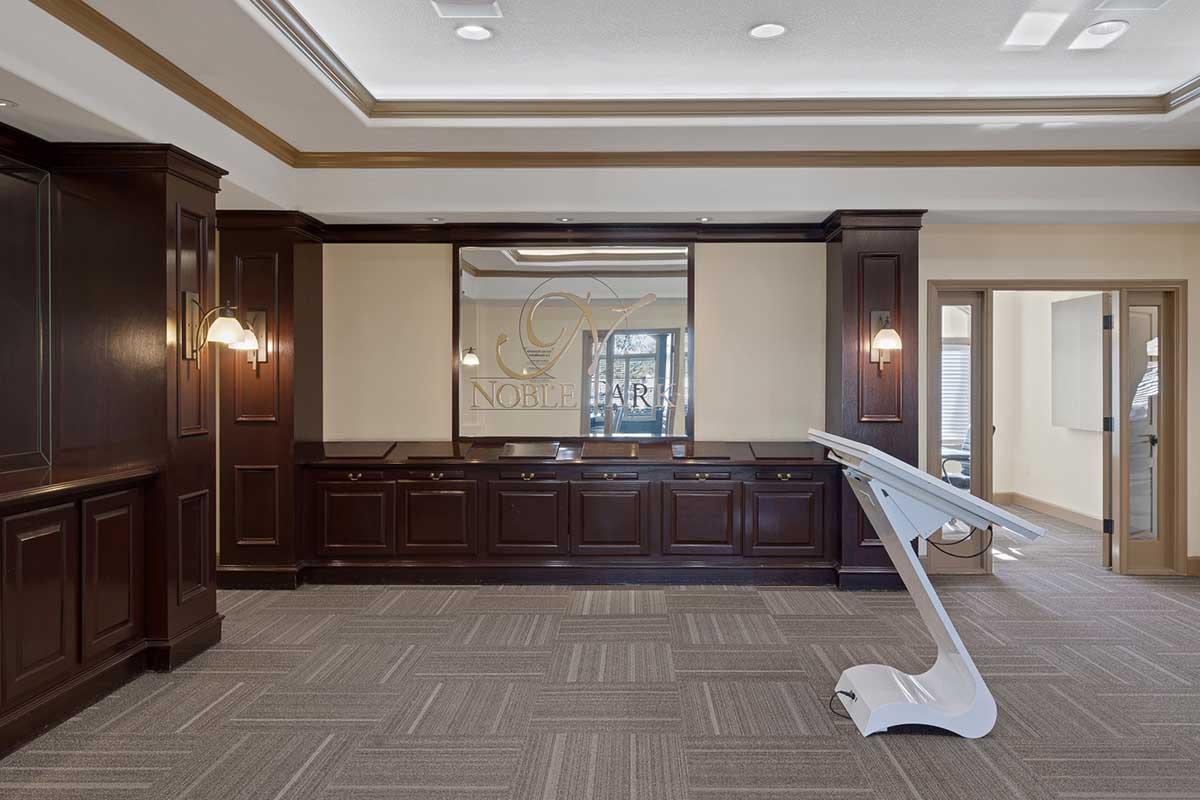 Interior of a modern office space featuring dark wood paneling, a reception desk with a large mirror, and an elegant lighting fixture. The floor is covered with gray carpet tiles, and a white, sleek stand is positioned in the foreground. A doorway leads to another room, creating a professional and welcoming atmosphere.