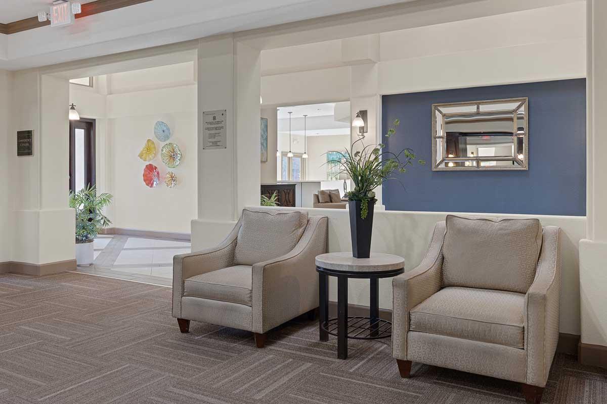 A cozy lobby area featuring two light-colored armchairs flanking a small round table. A decorative plant sits on the table, and in the background, colorful wall art can be seen. Soft lighting and neutral-toned walls create a warm, inviting atmosphere.