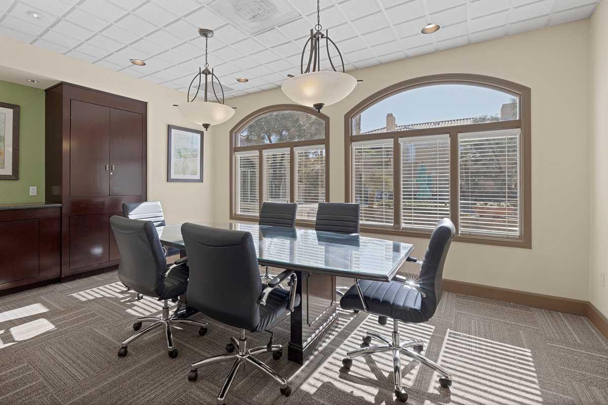 A well-lit conference room featuring a large glass table surrounded by black leather chairs. The room has two pendant lights hanging from the ceiling and large windows with blinds, allowing natural light to fill the space. Neutral-colored walls and a patterned carpet create a professional atmosphere.