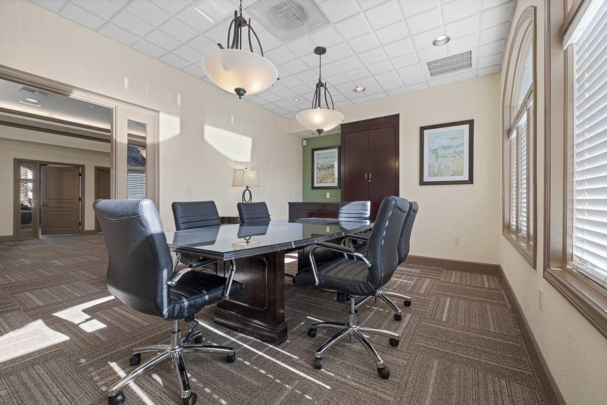 A modern conference room featuring a large glass table surrounded by black leather executive chairs. The space has neutral-colored walls, multiple light fixtures, and large windows with blinds that let in natural light. A dark wood cabinet and framed artwork are visible in the background.