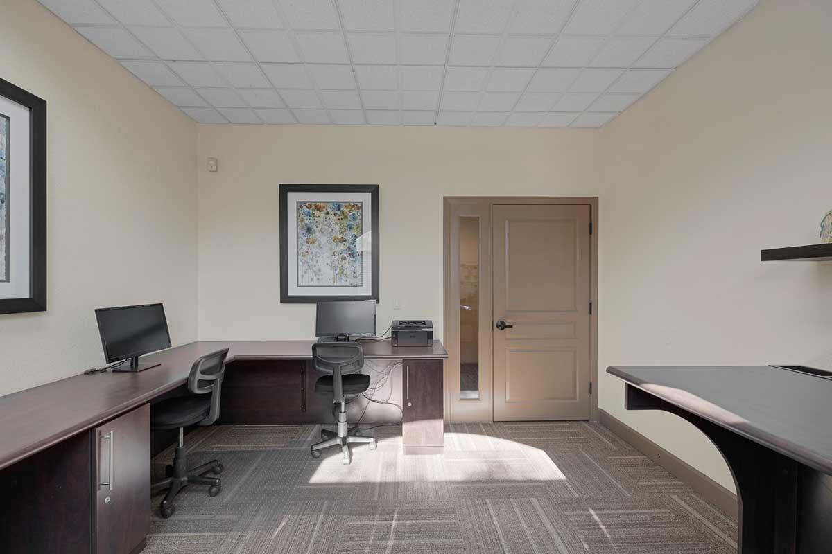 A modern office space featuring two desks with black chairs, a computer on one desk, and a printer in the corner. The room has light-colored walls, a textured ceiling, and a decorative framed artwork on one wall. Natural light fills the room from a door leading to another space.